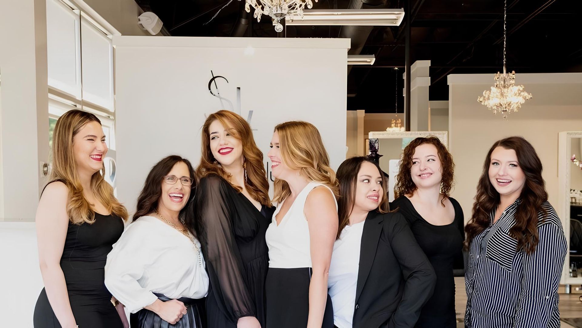 Seven standing hairstylists in a hair salon looking and smiling at each other.
