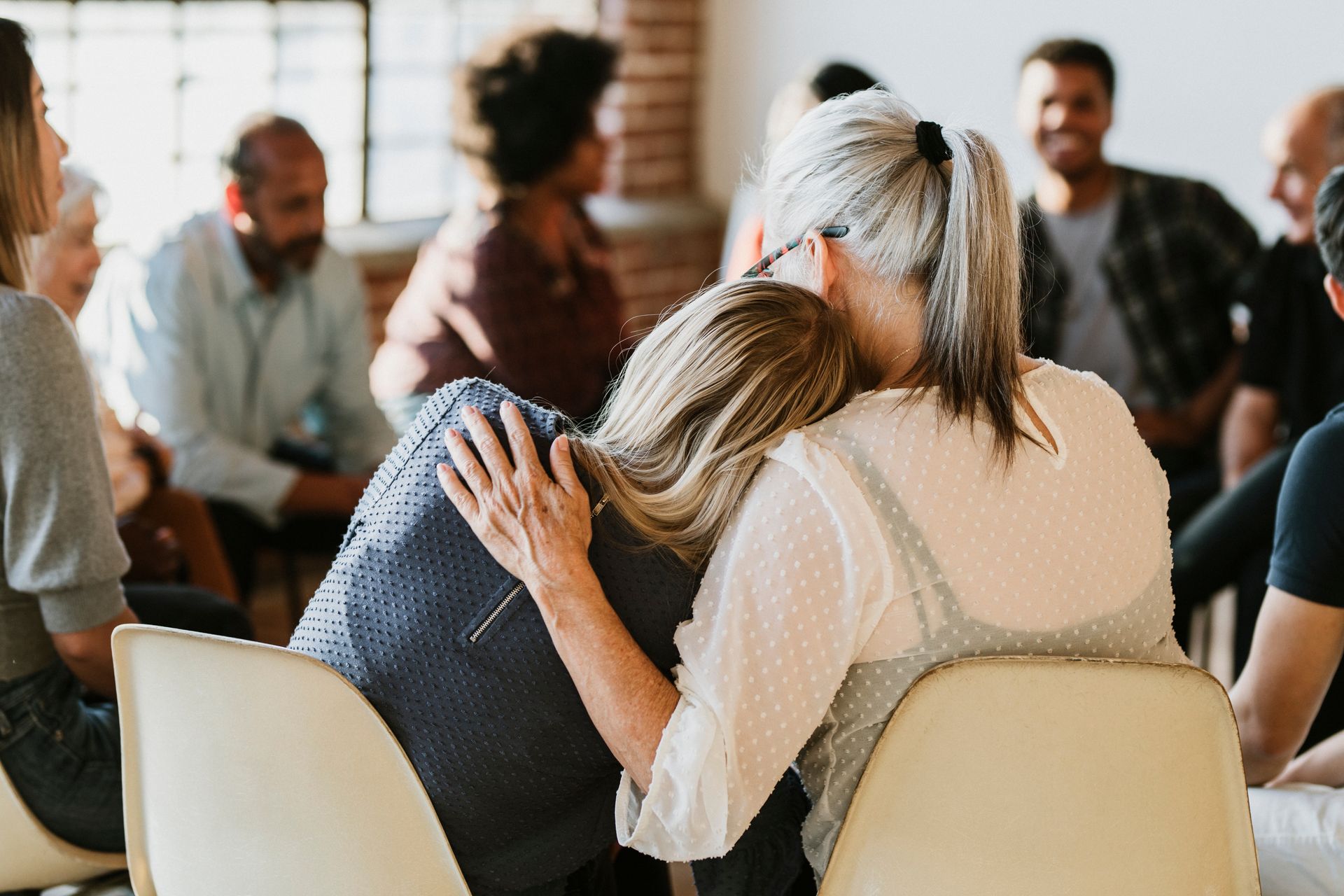 A woman is hugging another woman in a group of people sitting in chairs.