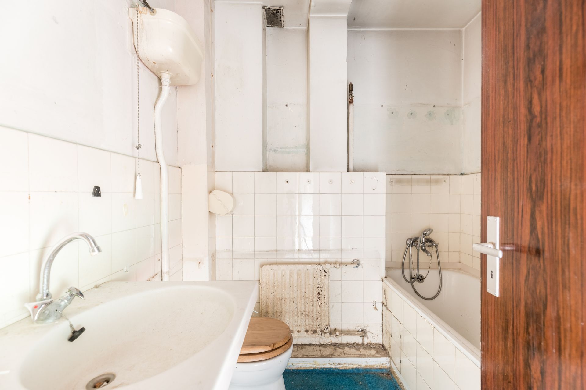 Run-down bathroom with a sink, toilet, tub, and peeling white tiles. Brown wooden door.