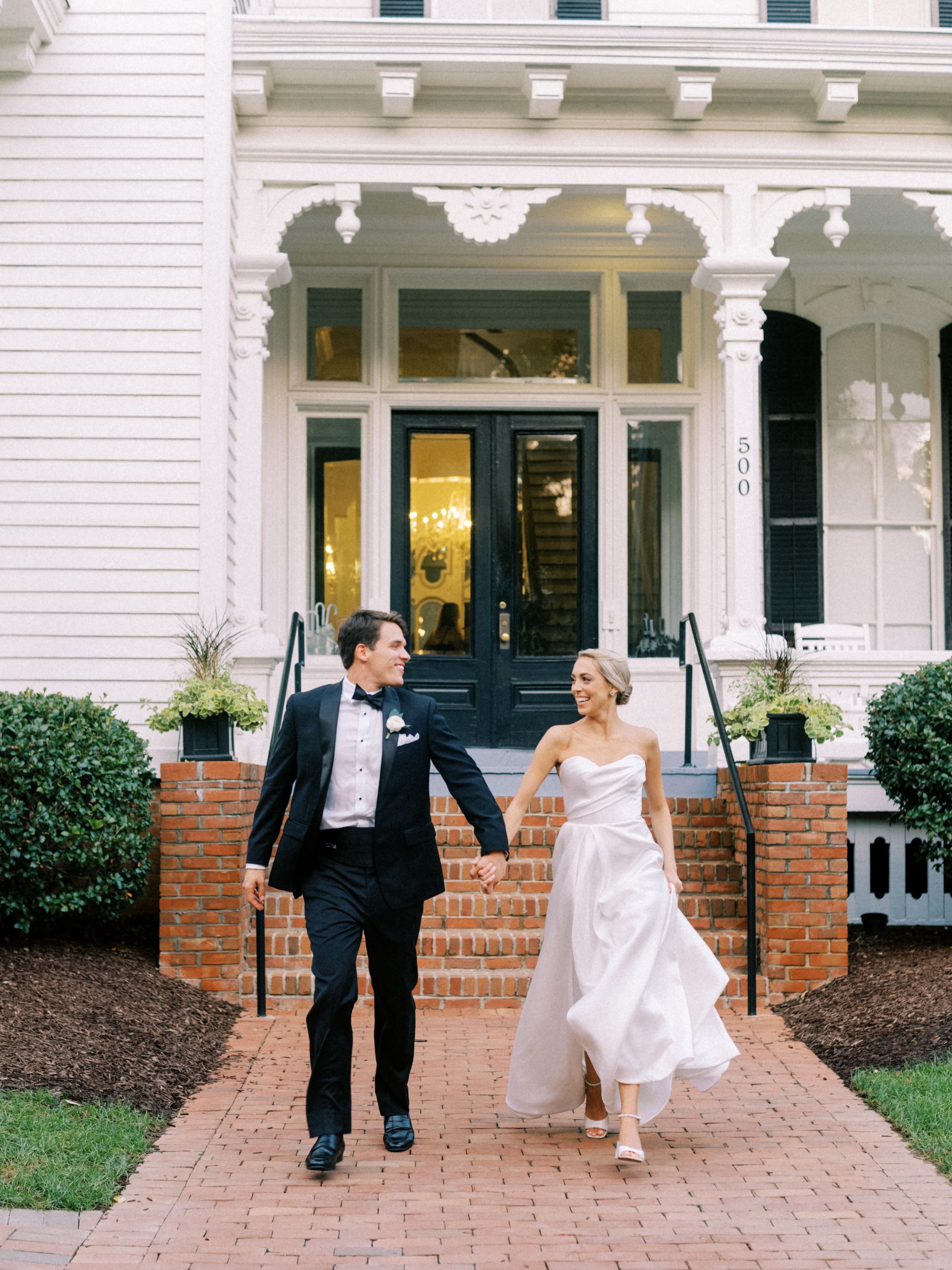 A couple in wedding attire walks hand-in-hand down a brick path toward the camera in front of a white historic home.