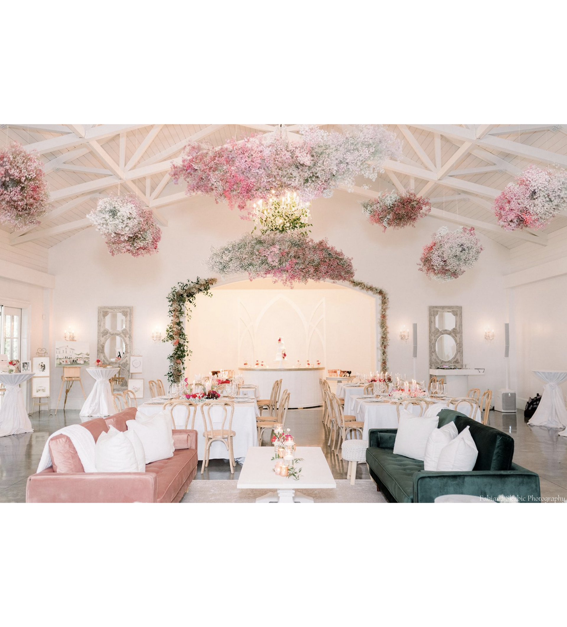 A large room with tables and chairs and pink and white babies breath flowers hanging from the ceiling. There are pink and green velvet couches that create lounge areas for wedding guests to enjoy. 