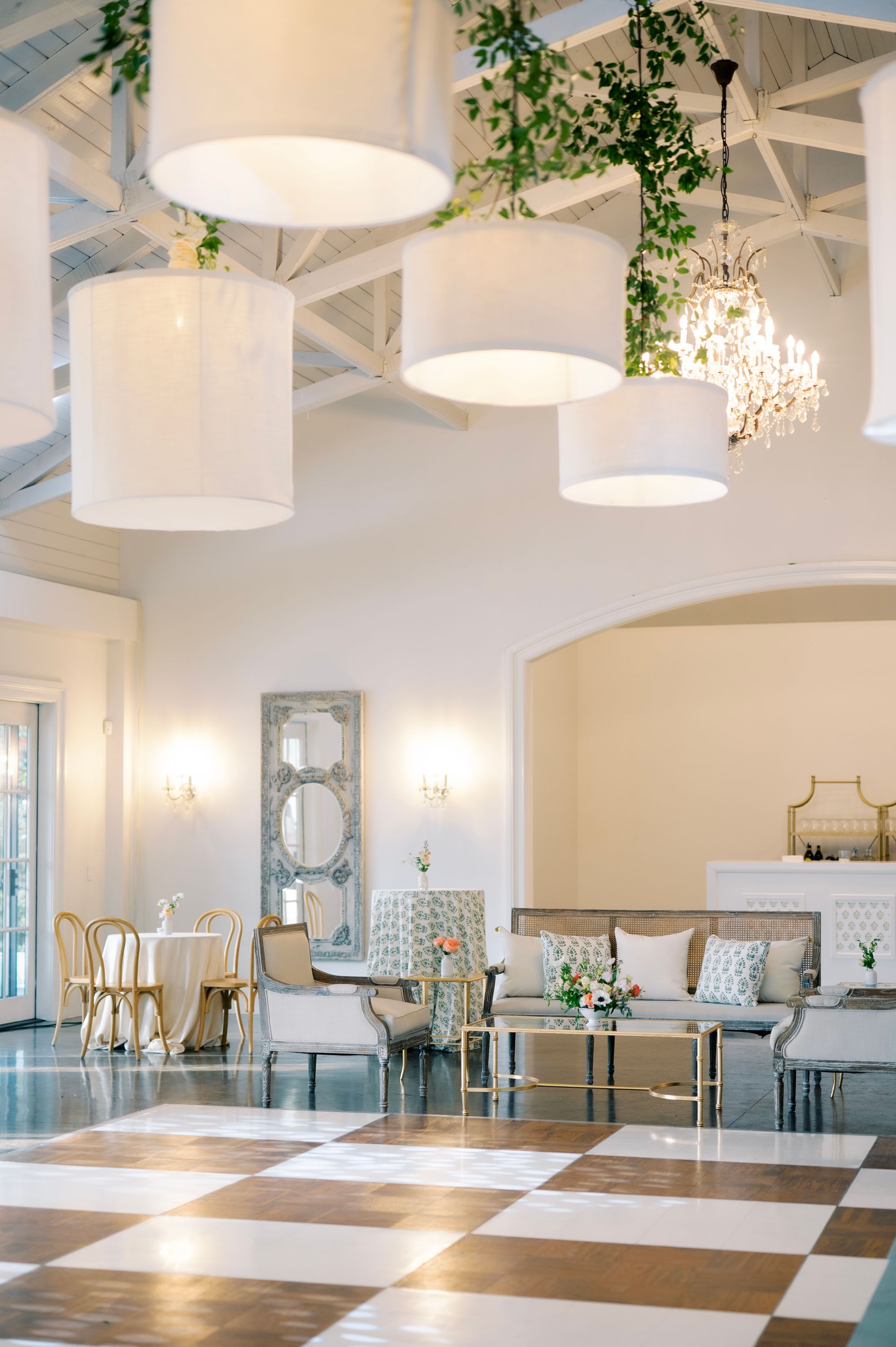 An elegant ballroom with a checkered dance floor, large hanging drum lights, greenery, and sophisticated lounge seating.