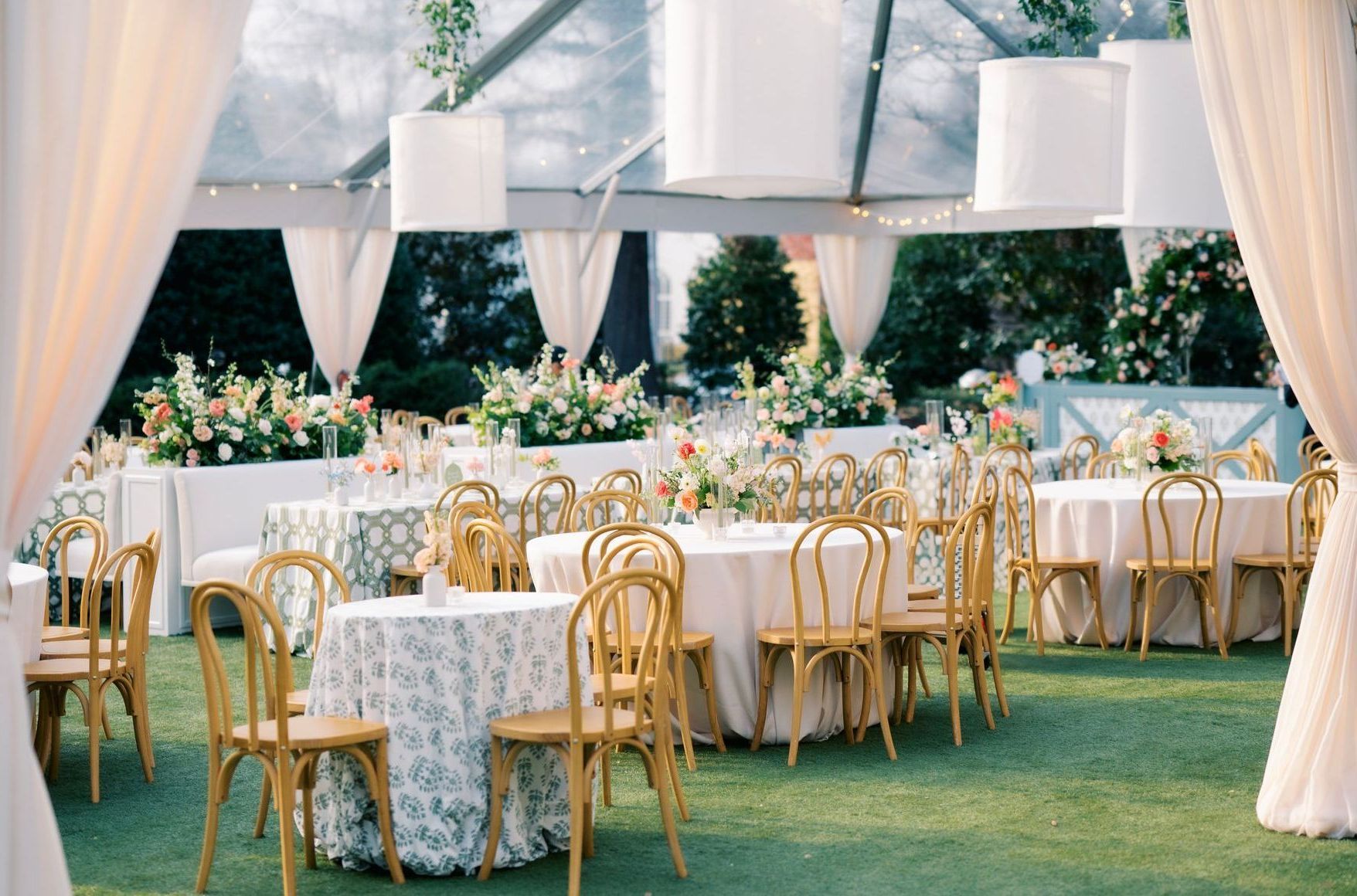 A garden wedding reception under a clear marquee tent with round tables, gold chairs, and floral arrangements.