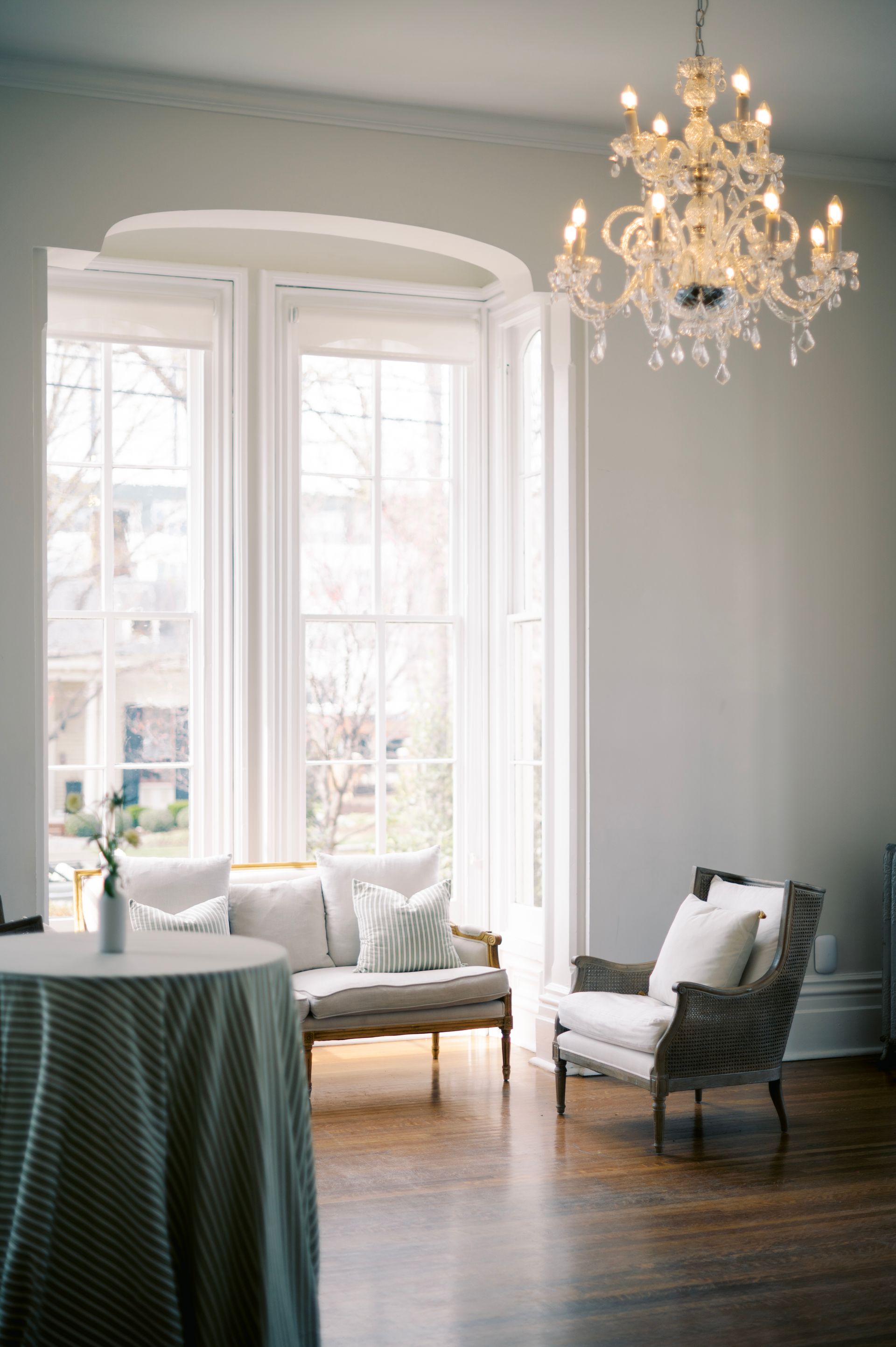 A bright, elegant room with a crystal chandelier, a white bay window, a small settee, and a patterned armchair.