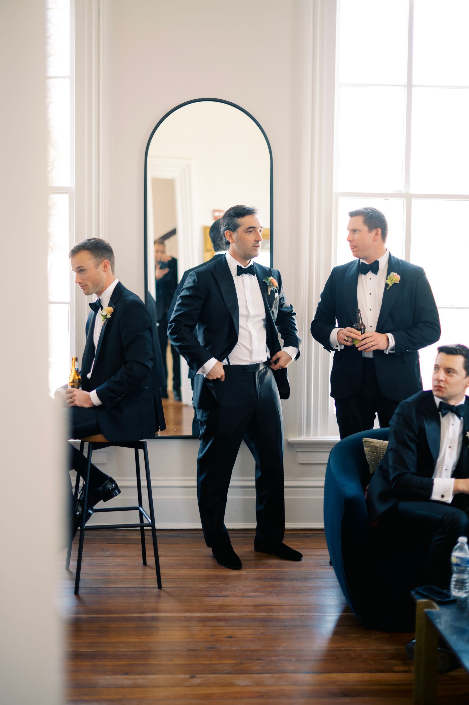 Four men in dark tuxedos gather in a bright, elegant room with high windows, a floor mirror, and hardwood floors.