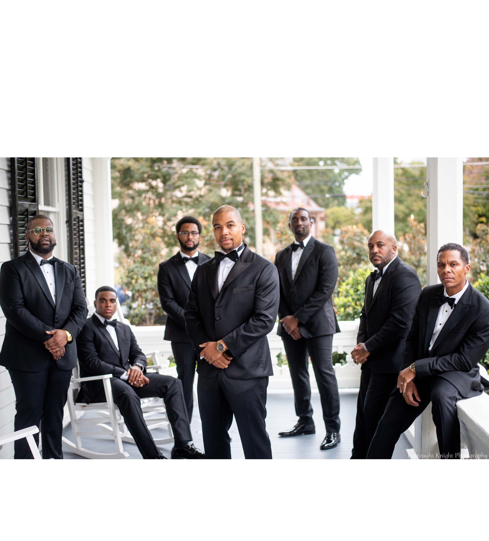 A group of men in black suits with bow ties are standing and sitting on a porch surrounding the groom. 