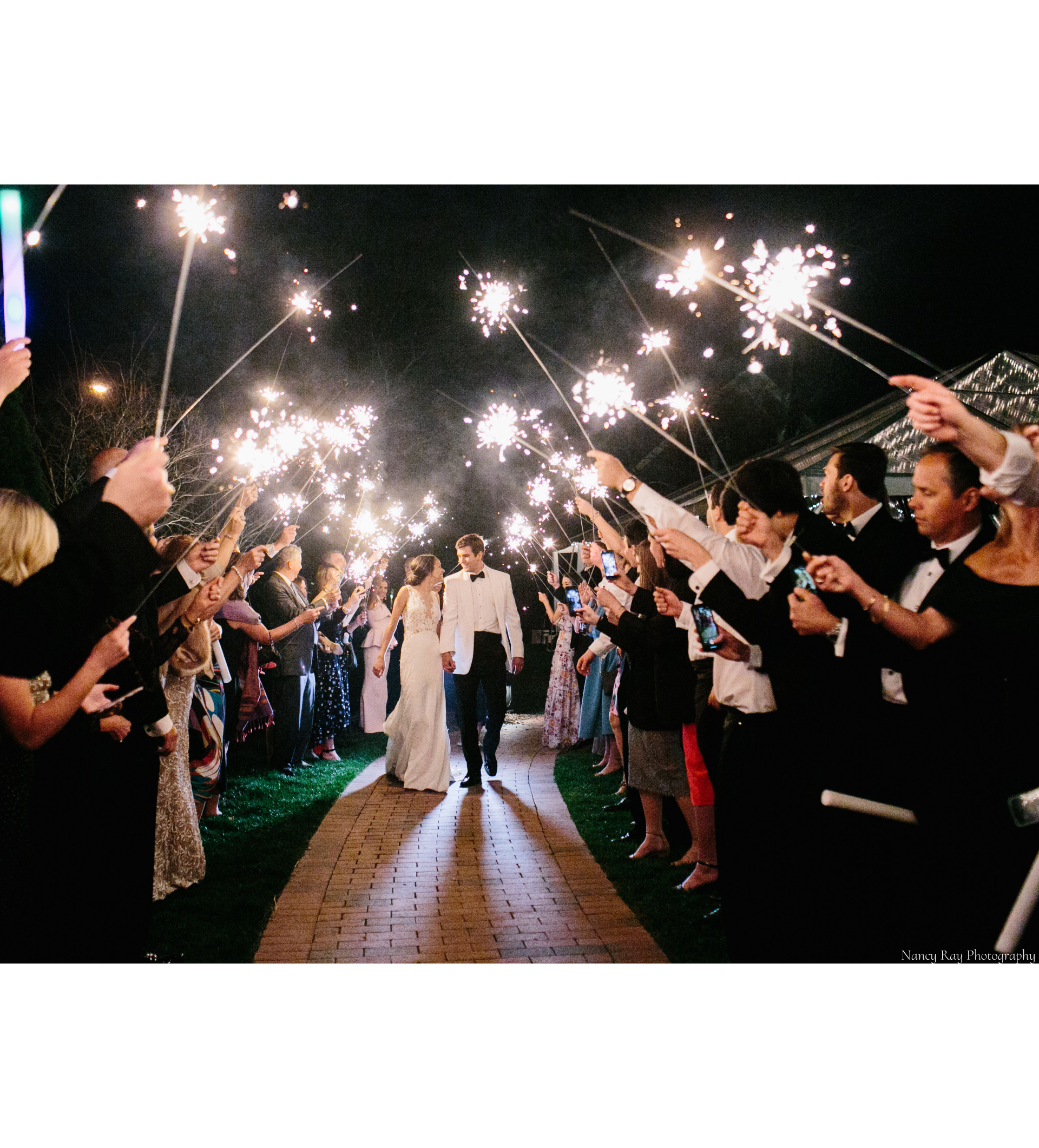 A bride and groom are walking down a brick path through a tunnel of sparklers held by their wedding guests.