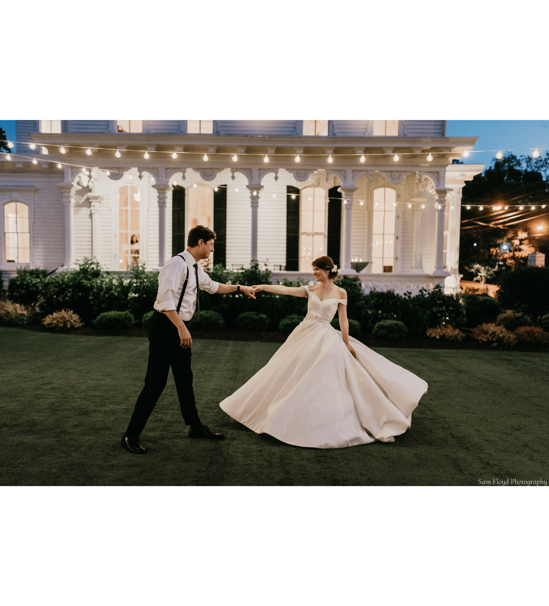 A bride and groom are dancing in front of a large white house during sunset, on the lawn with market lights above.