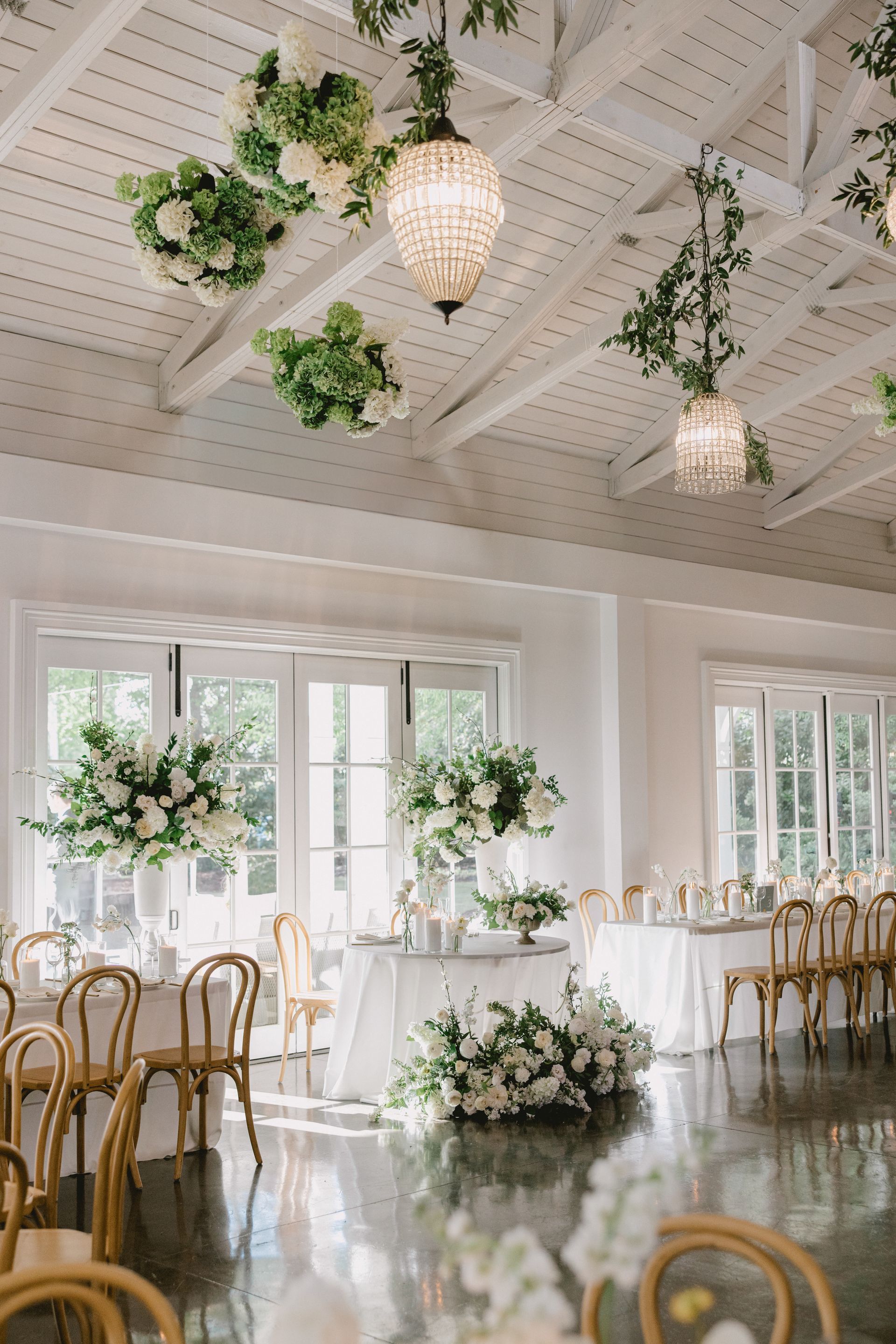 Elegant wedding reception venue with white draped ceilings, hanging floral arrangements, chandeliers, and dining tables.