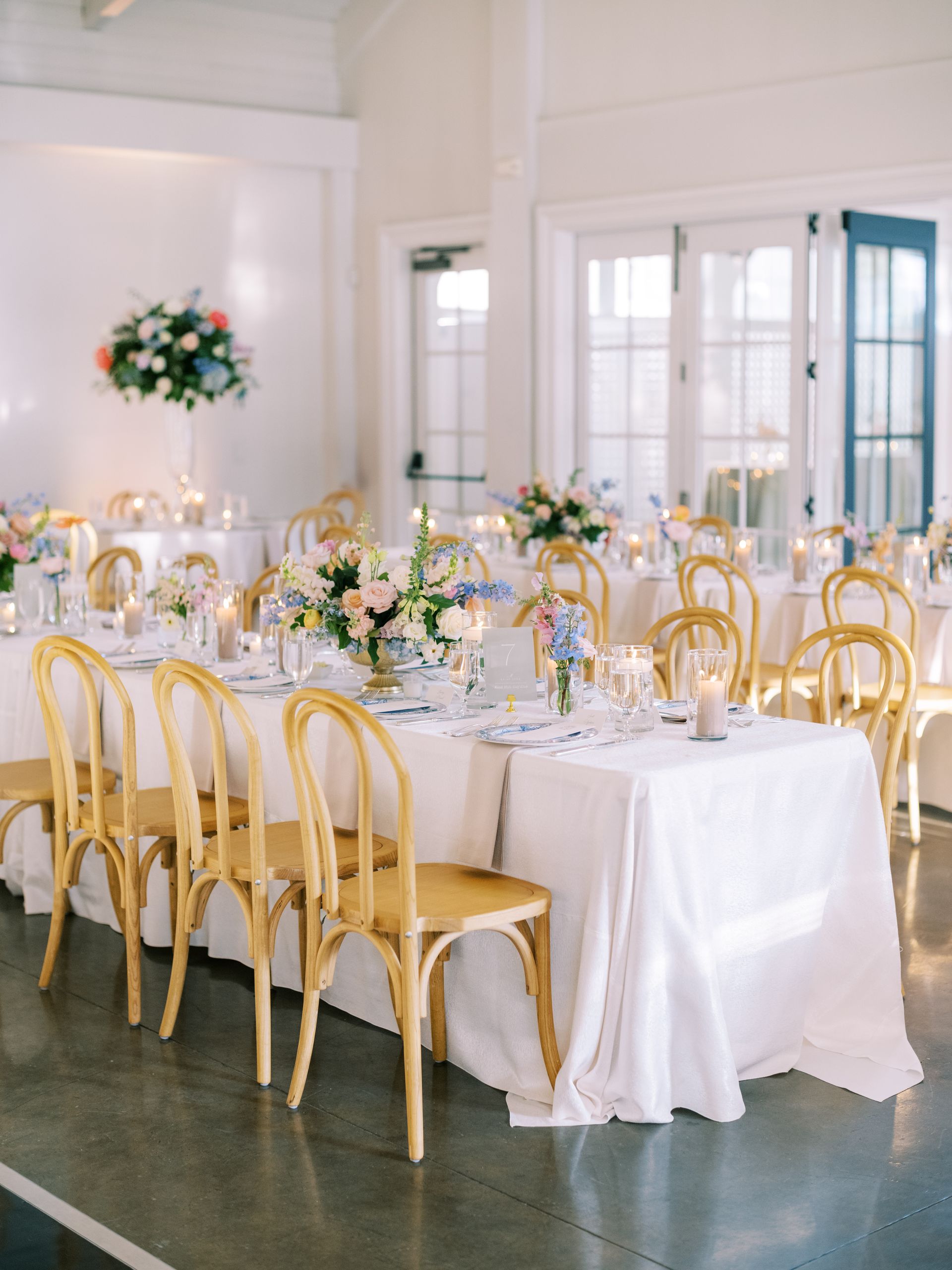 A large white room filled with tables that are dressed for a wedding reception dinner with light brown chairs around for guests.