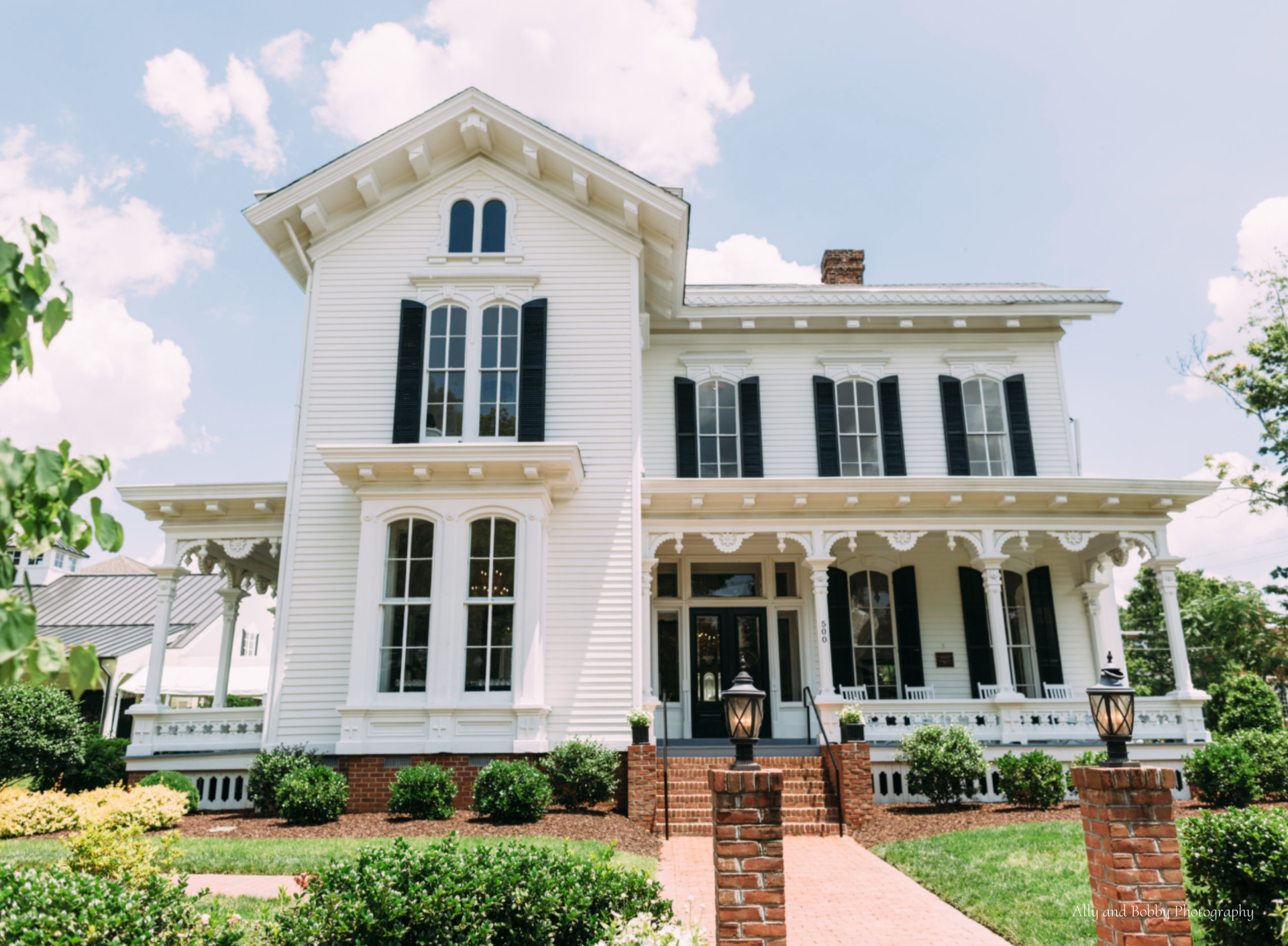 A large white house with black shutters, a large porch and brick stairs leading to the front door.