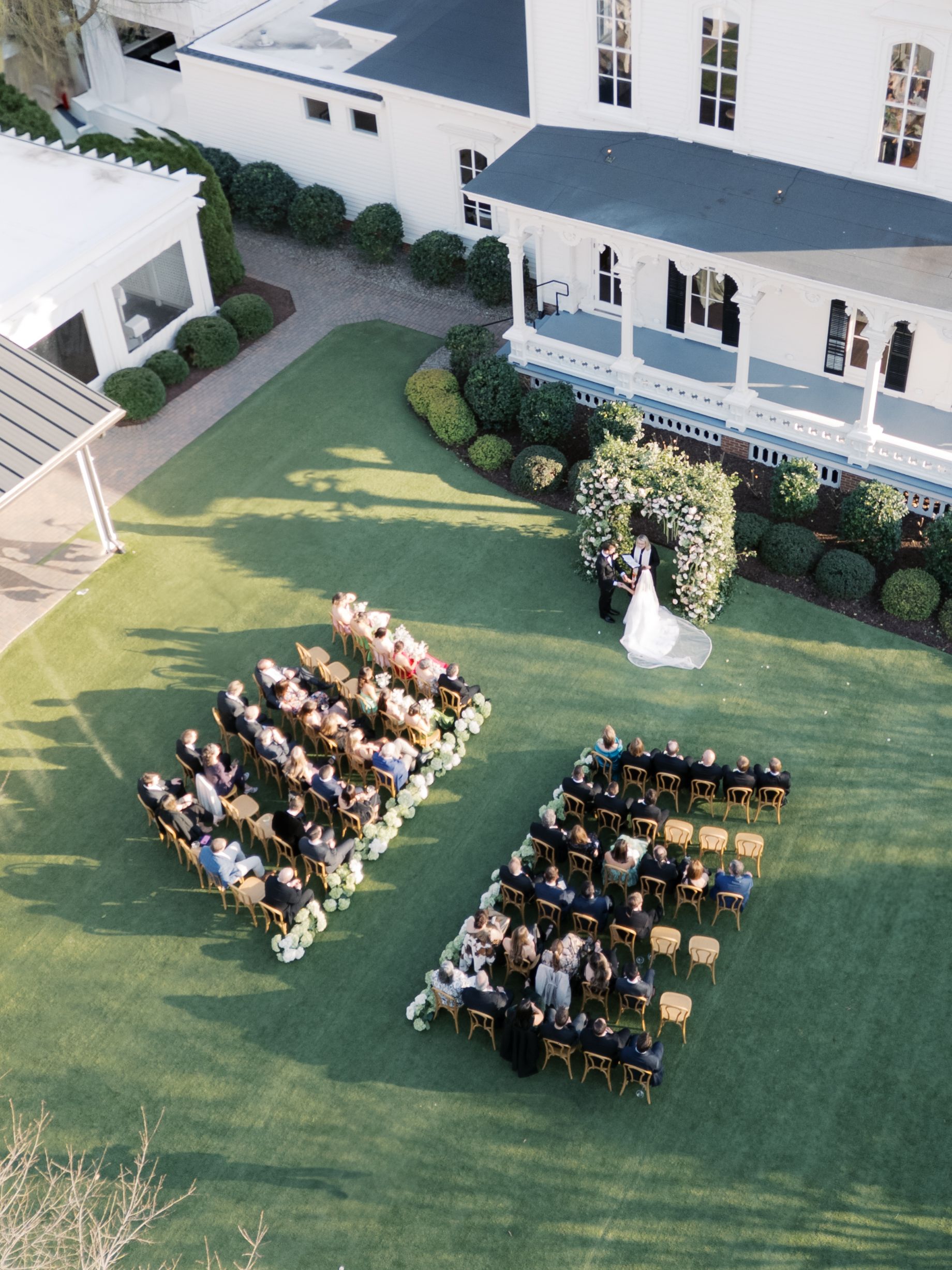 A clear-roofed event tent with tables, floral arrangements, and blue sofas, set on a lawn with hanging greenery.