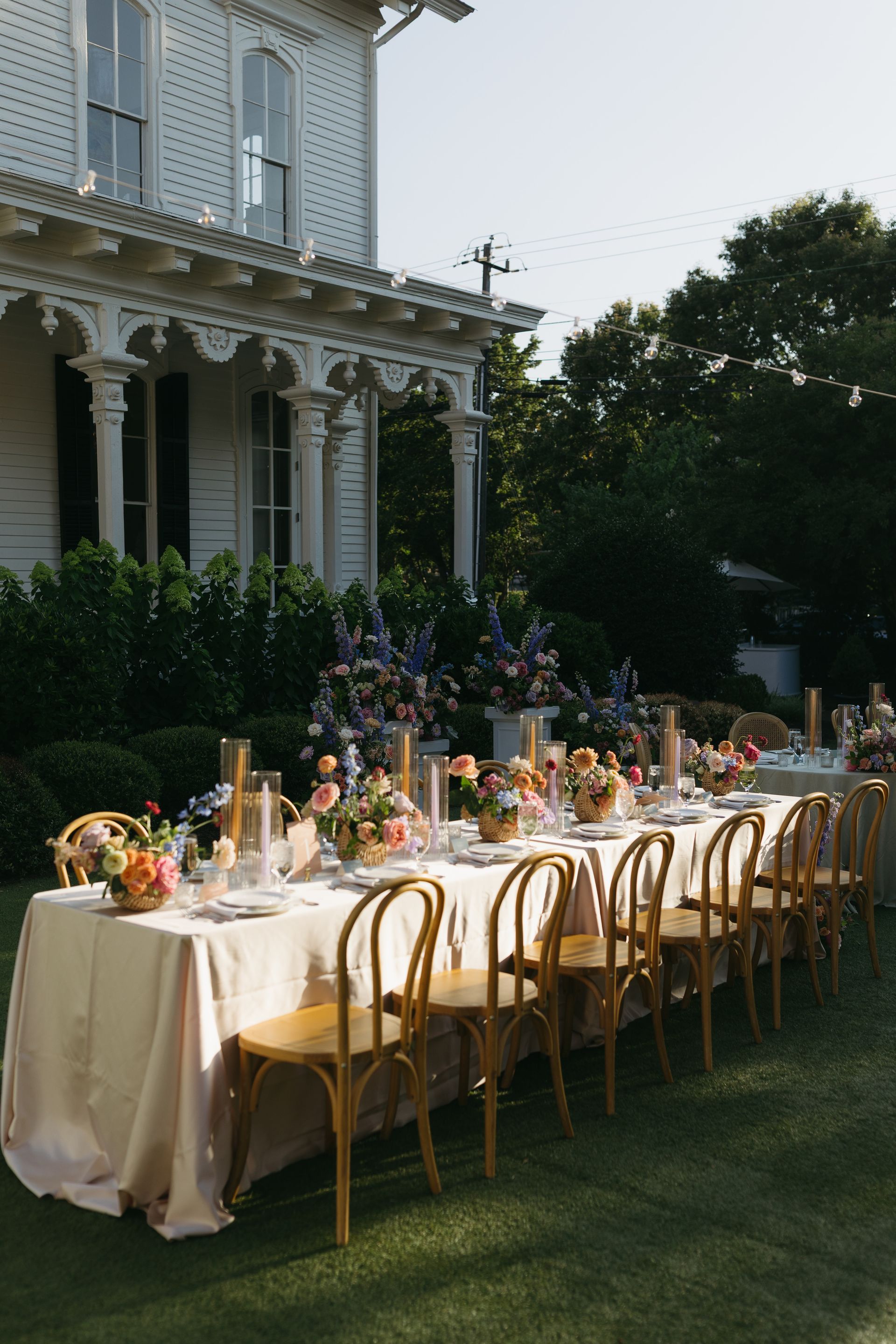 A long dining table set for an outdoor event on a lawn in front of a white historic house with a large porch.