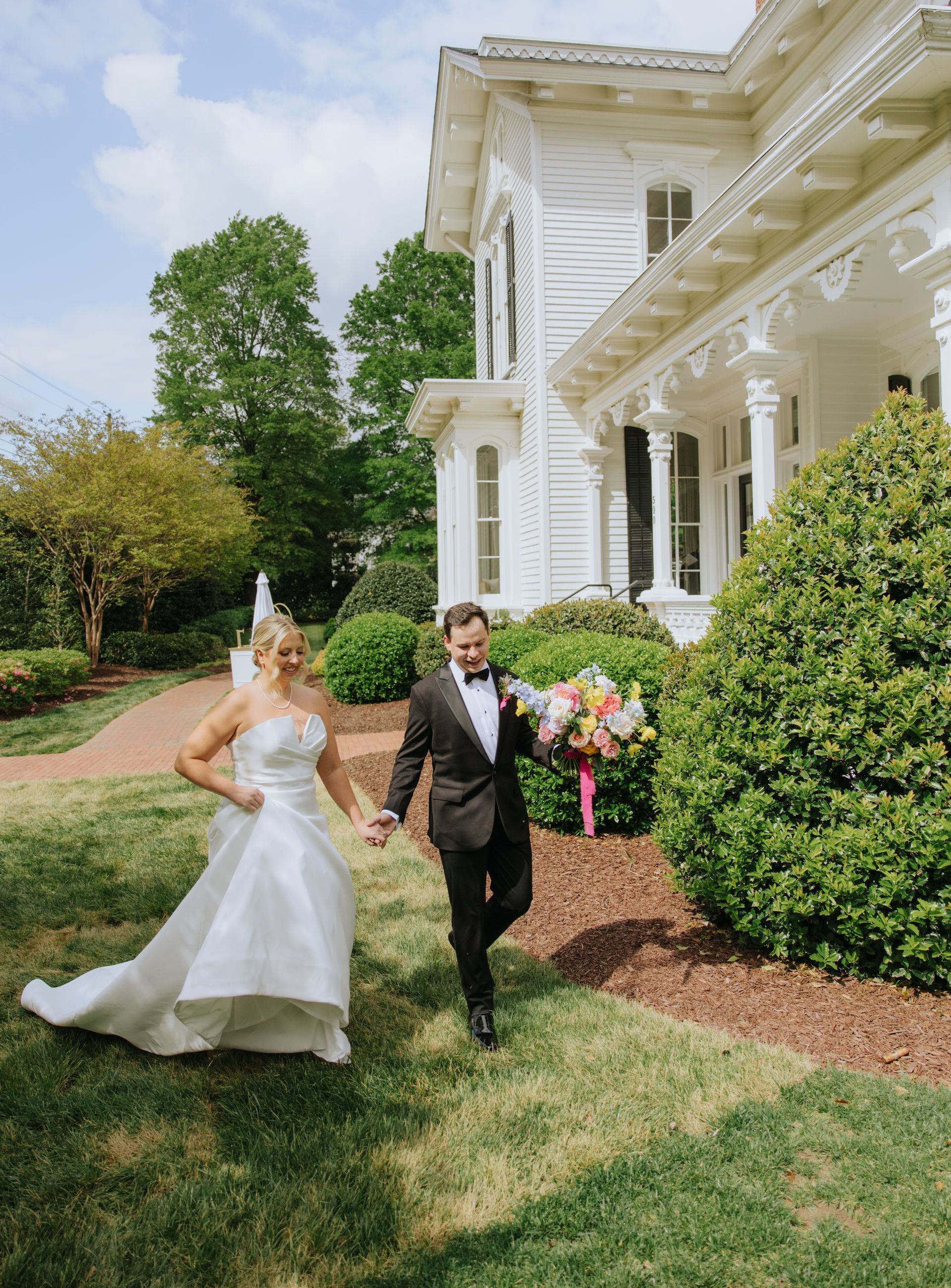 A bride and groom walk hand-in-hand on a lawn in front of a white historic house, the bride holding a colorful bouquet.