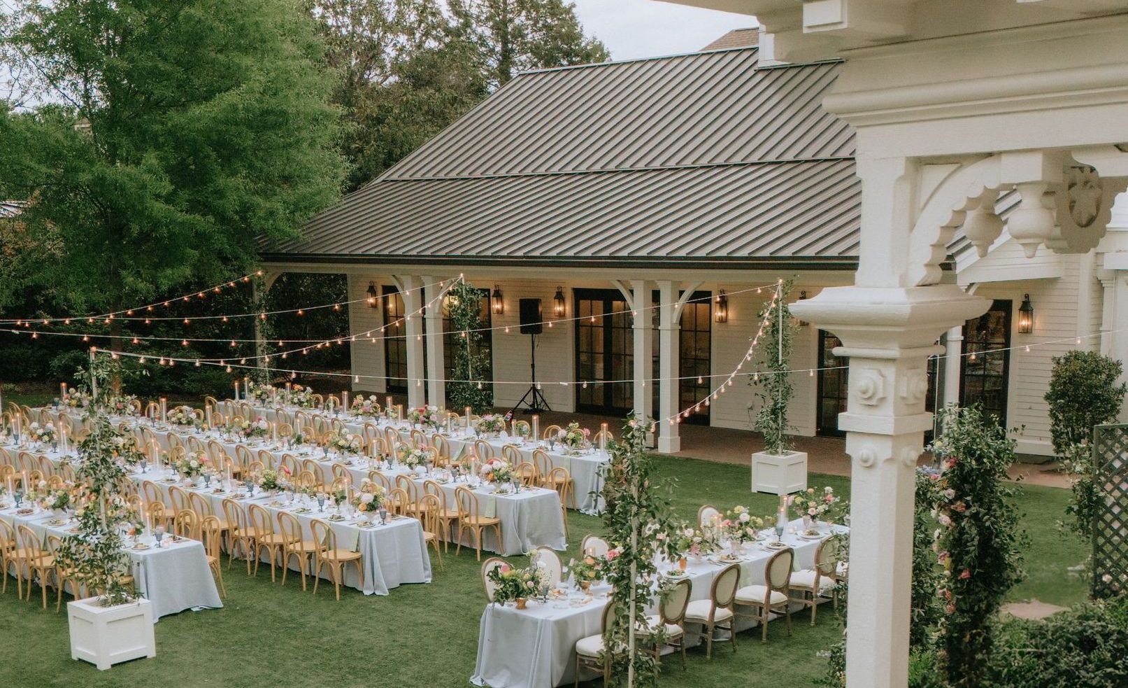 Outdoor wedding reception with long, white-clothed tables set on a grassy lawn in front of a white building with string lights.