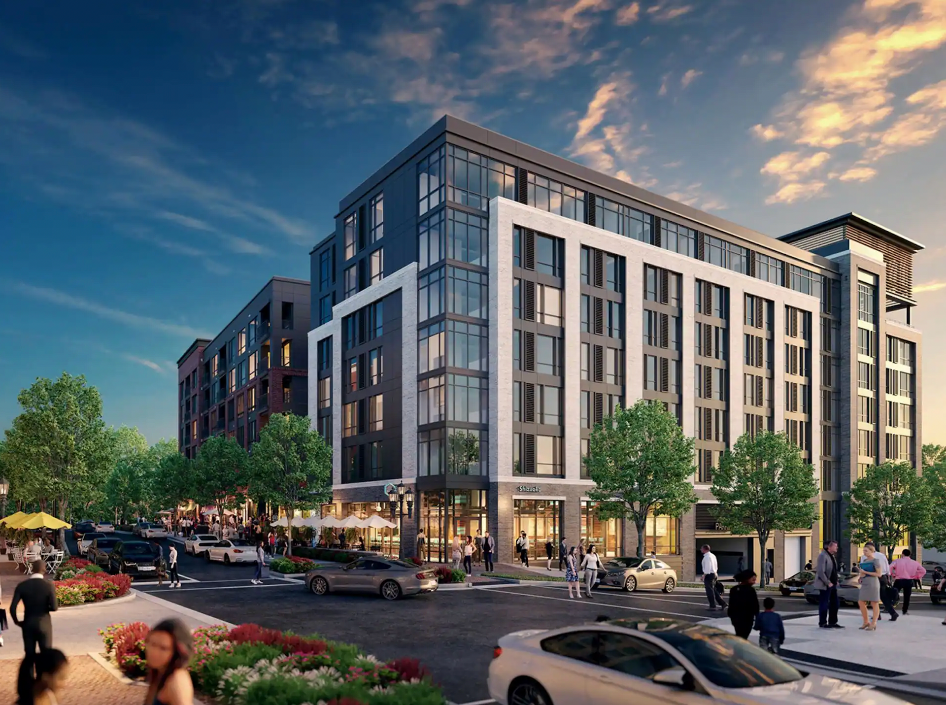 A modern multi-story apartment complex at sunset, featuring a ground-level retail area and pedestrians on a city street.