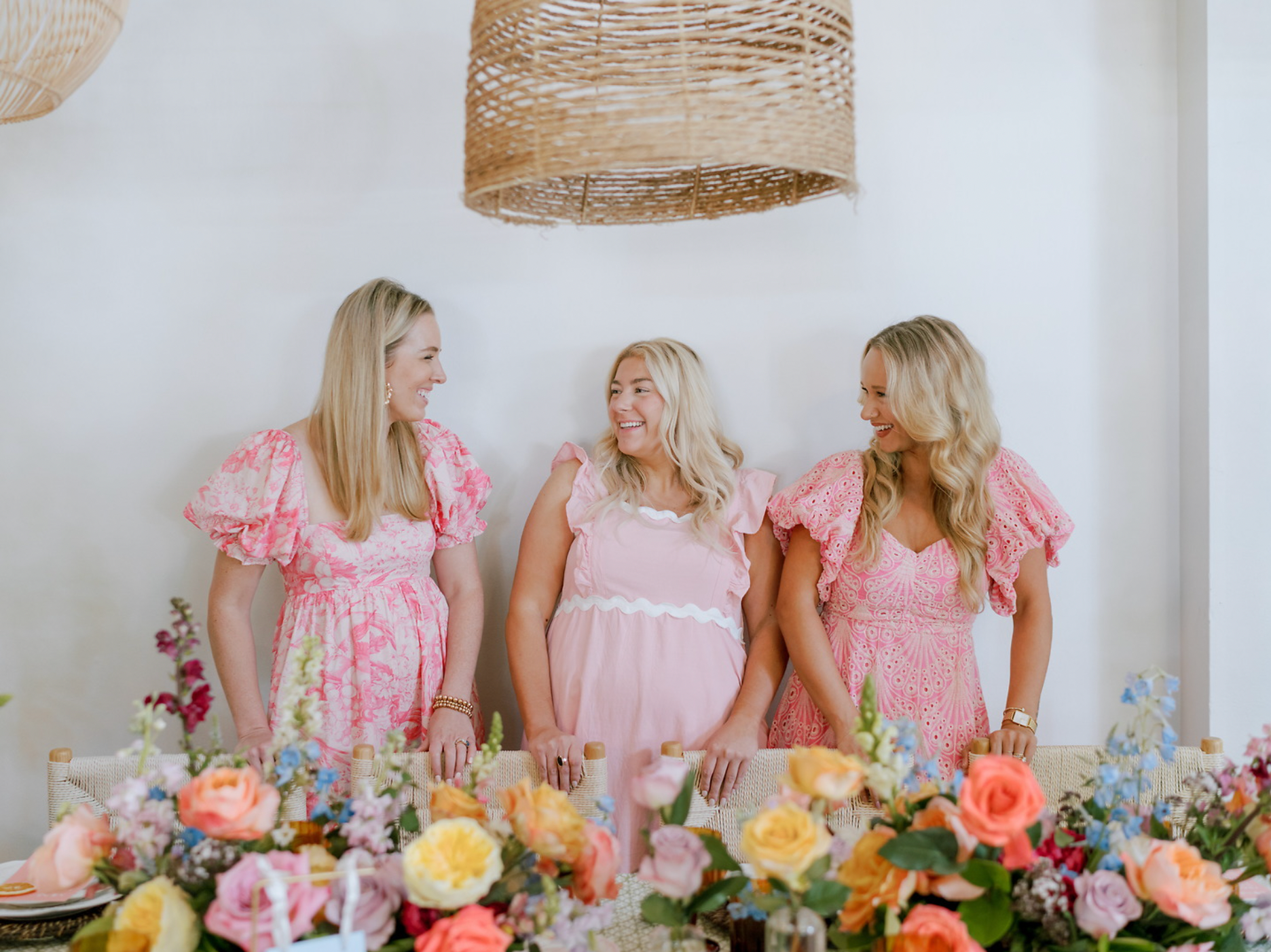 Three women in pink dresses are standing next to a table with yellow, orange and pink lush flowers.