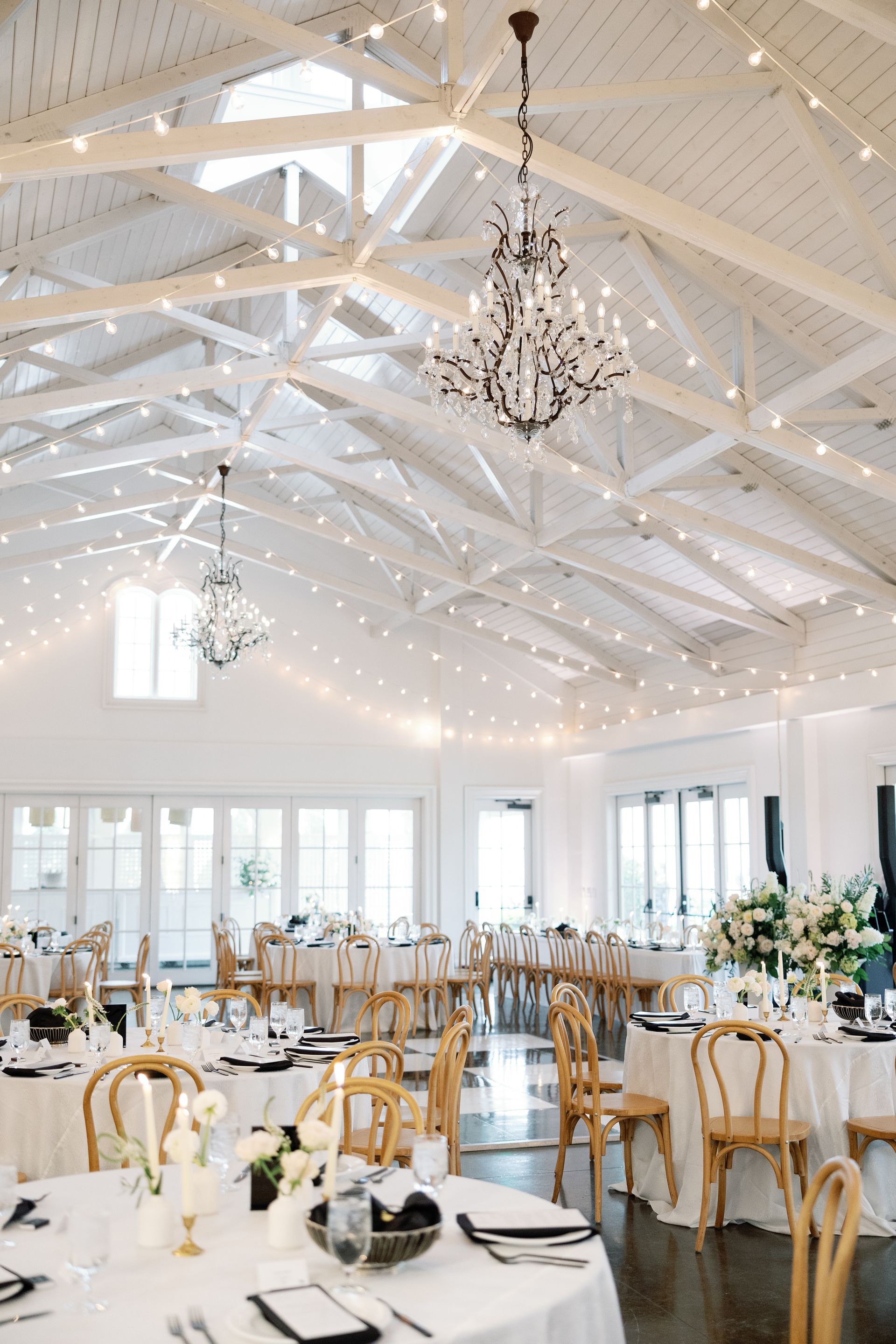 A bright, elegant event hall with a checkerboard dance floor, hanging drum pendants, greenery, and lounge seating.