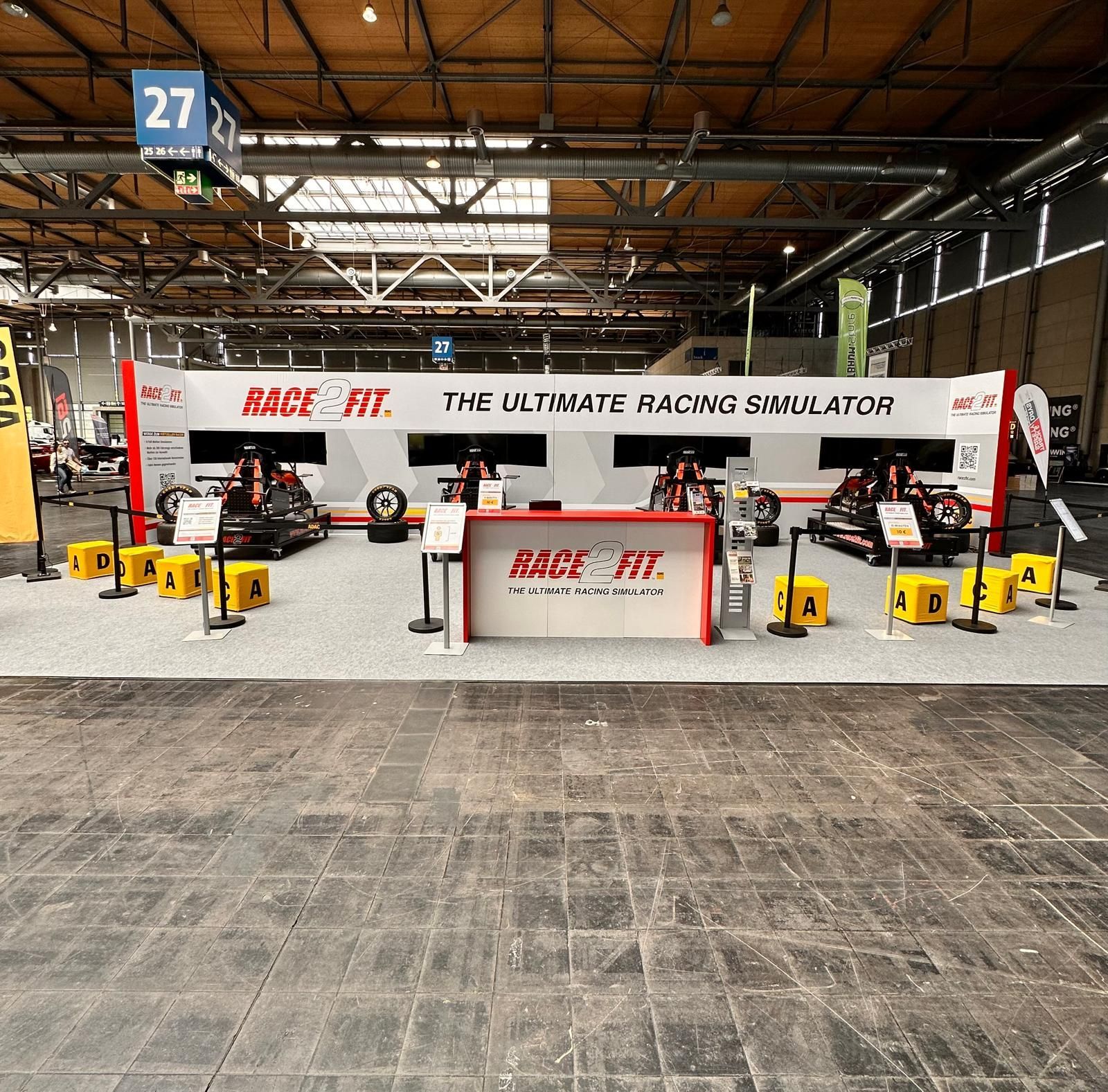 Stand des Rennsimulators RACE 1F in einer Indoor-Messehalle, mit weißen Displays und gelber Ausrüstung unter einer hohen Lagerhallendecke