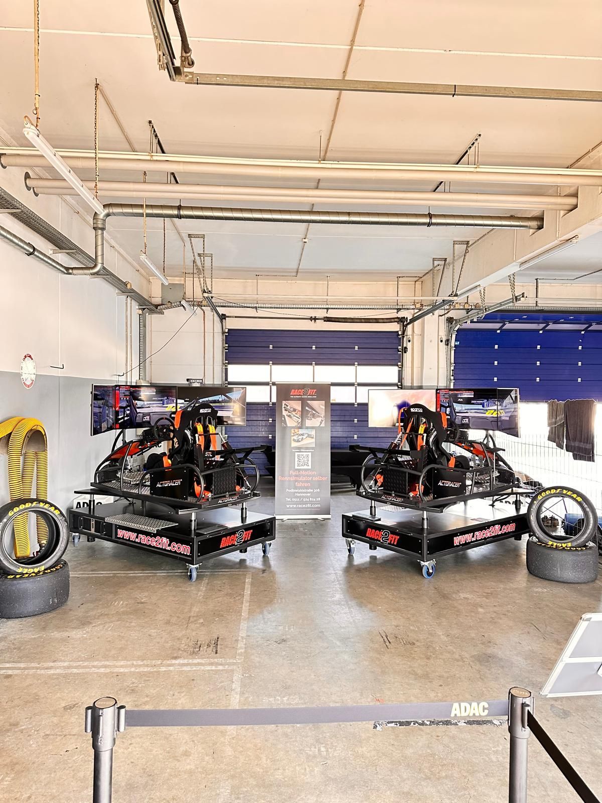 Renngarage mit zwei schwarzen Rennwagen, Reifen und offenen Toren unter hellem Deckenlicht