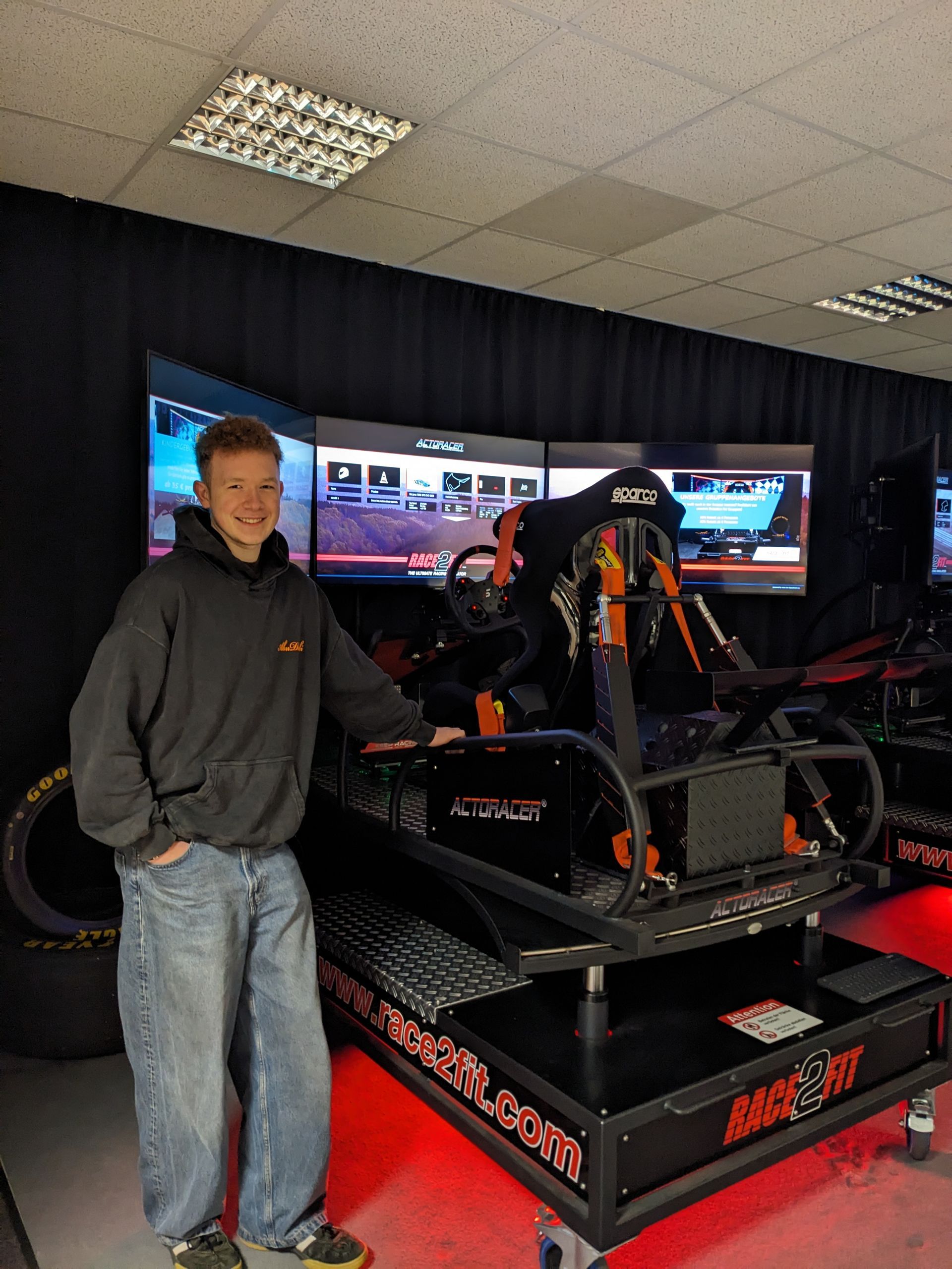 Eine Person steht neben einem Rennsimulator-Kart in einem Ausstellungsraum mit dunklen Vorhängen und Monitoren.