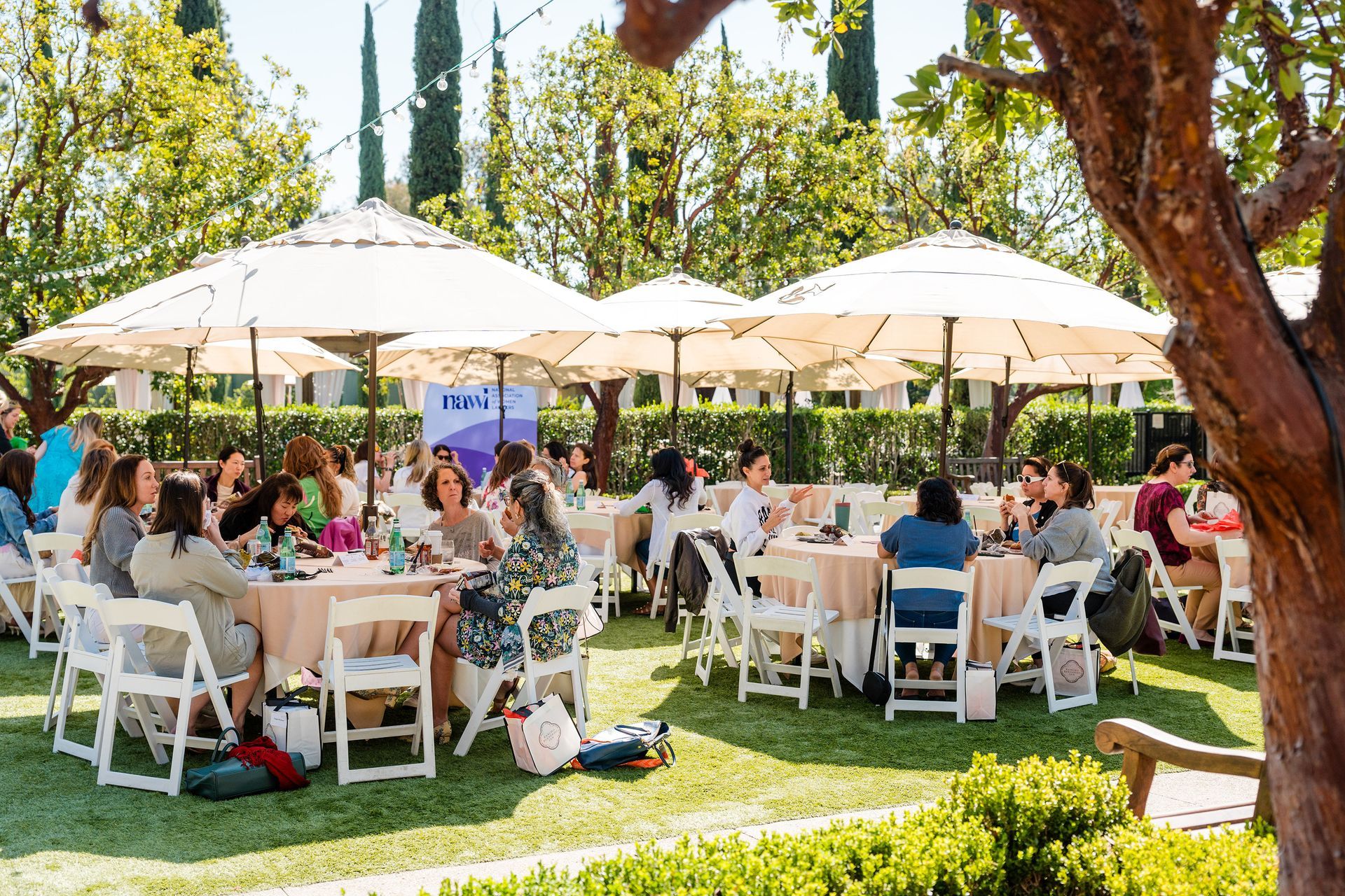 Participants sit at tables with umbrellas on a lawn.