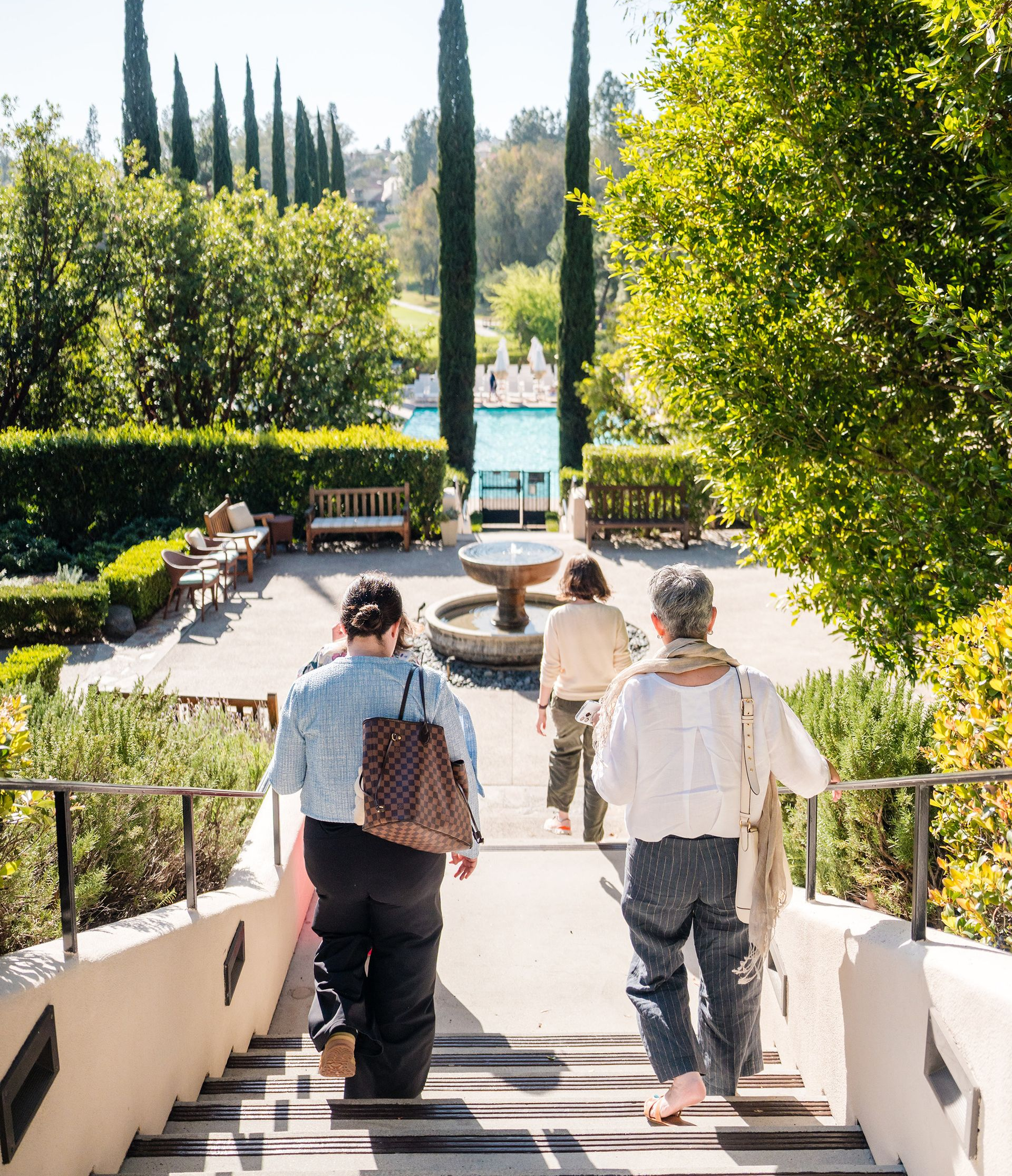 Women walking down stairs to a garden with trees and pool.