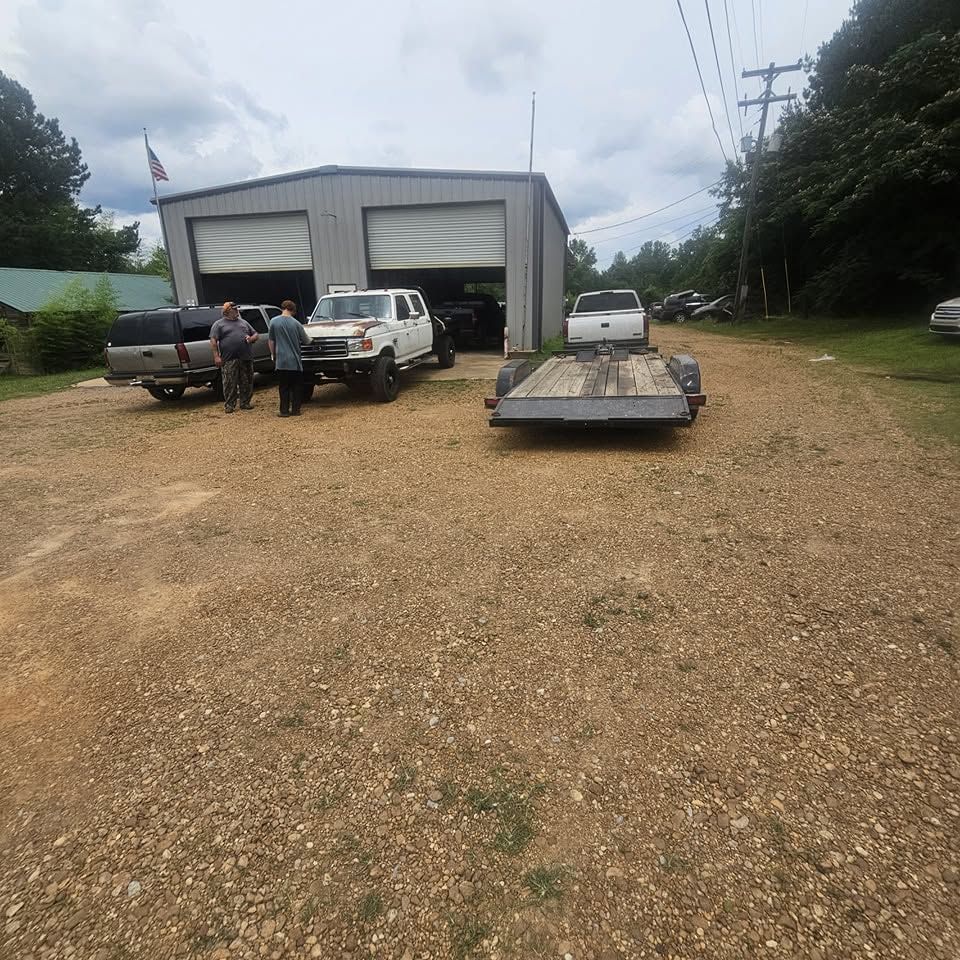 Gravel lot outside a warehouse with pickup trucks, a trailer, and two people standing near the vehicles.