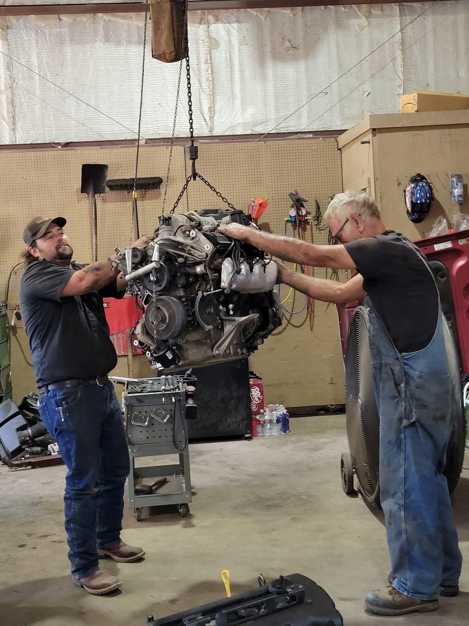 Two men in a workshop hoist a large engine with a chain hoist.