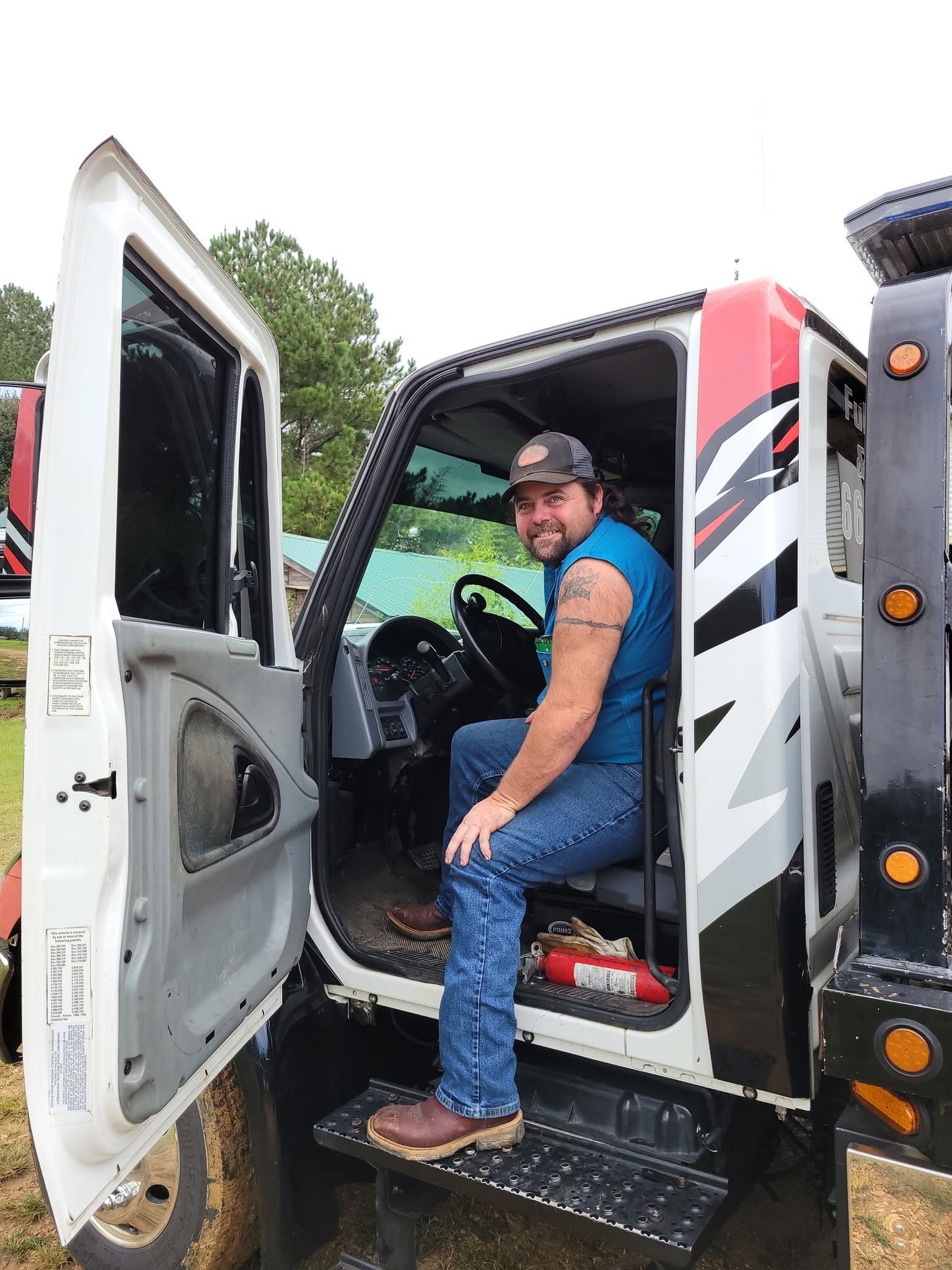 Person seated in the cab of a white truck with the door open, wearing a blue shirt and cap.