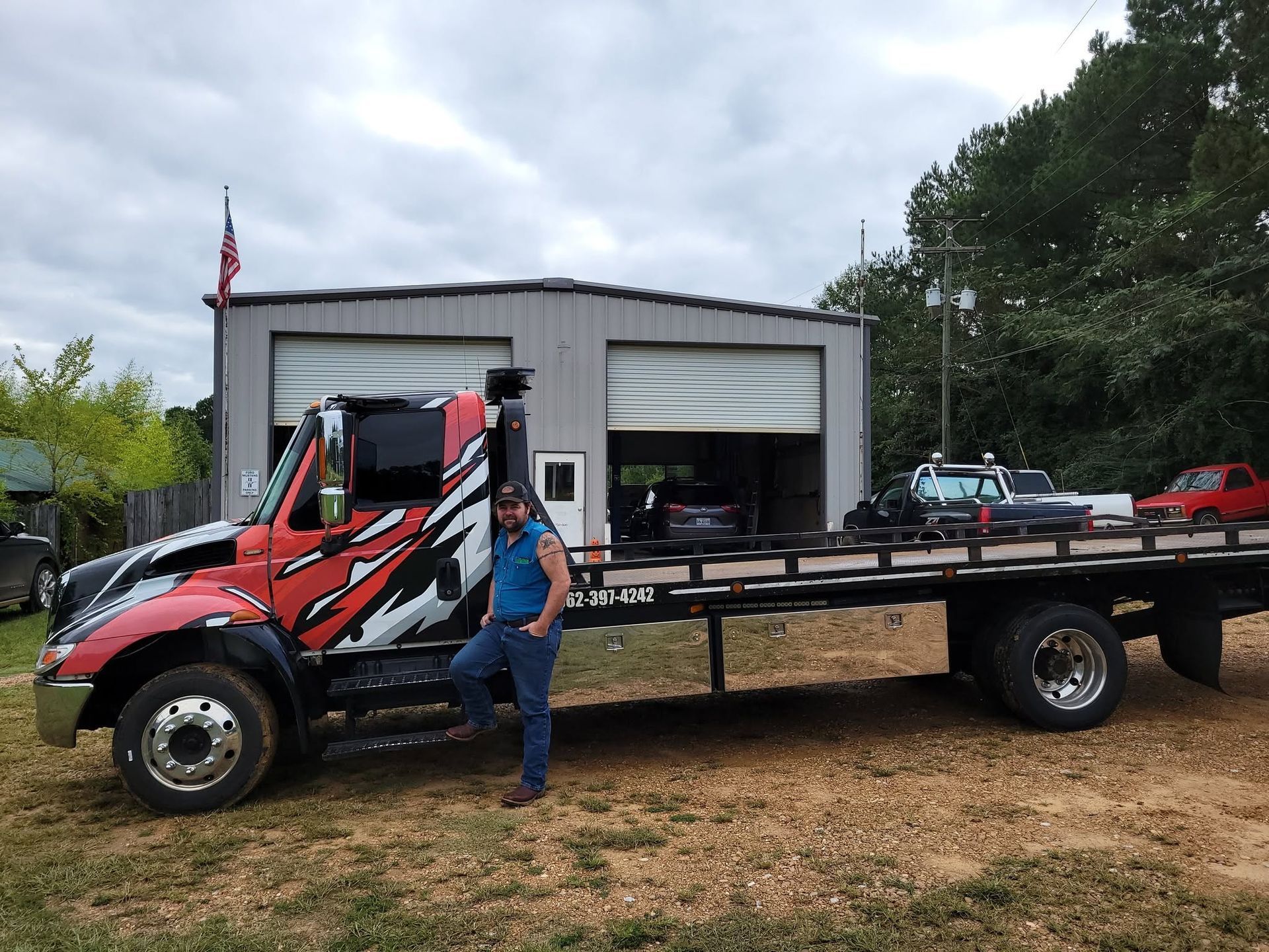 Red tow truck with flatbed parked outside a garage, with a person standing beside it.