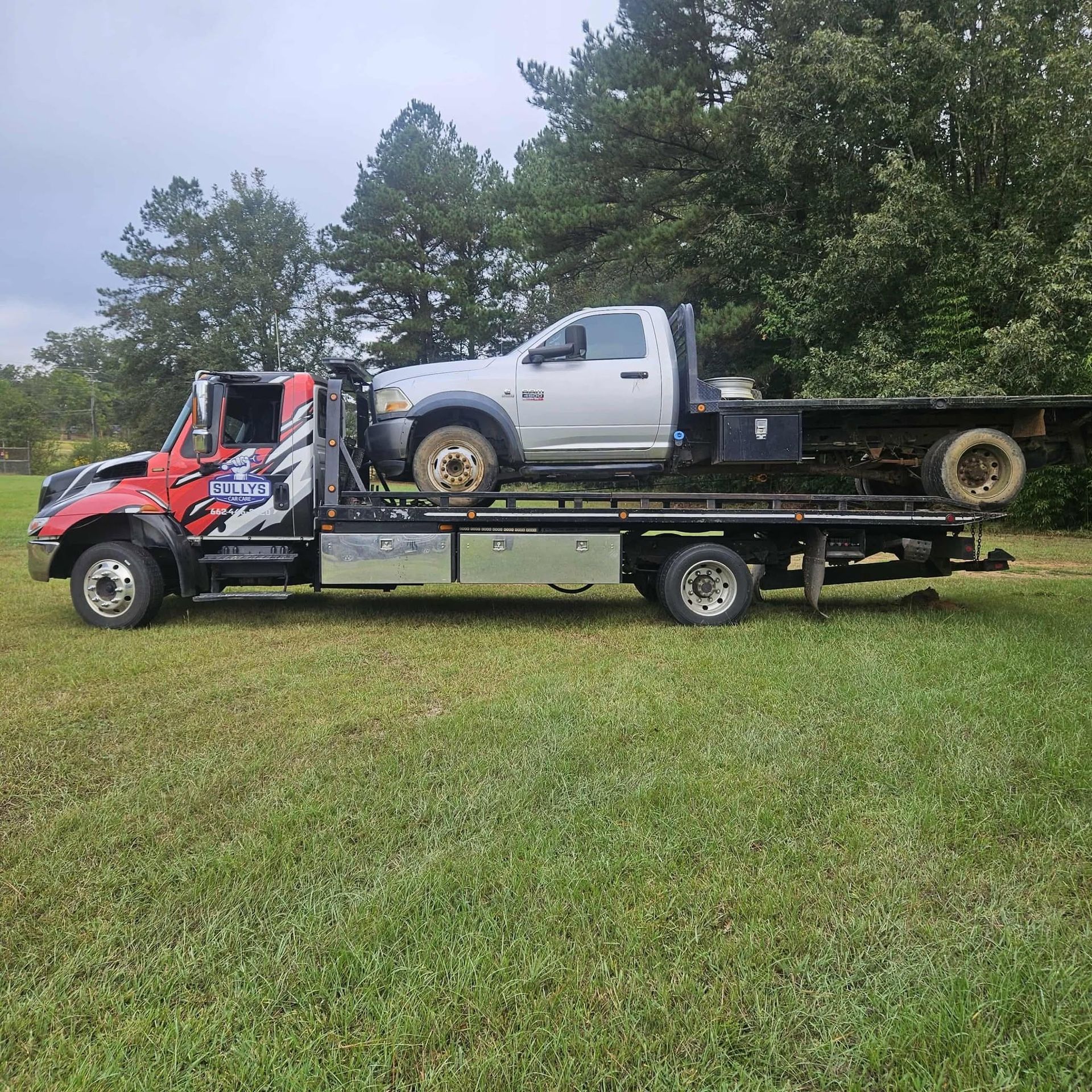Red tow truck carrying a white pickup on its flatbed in a grassy field with trees behind it