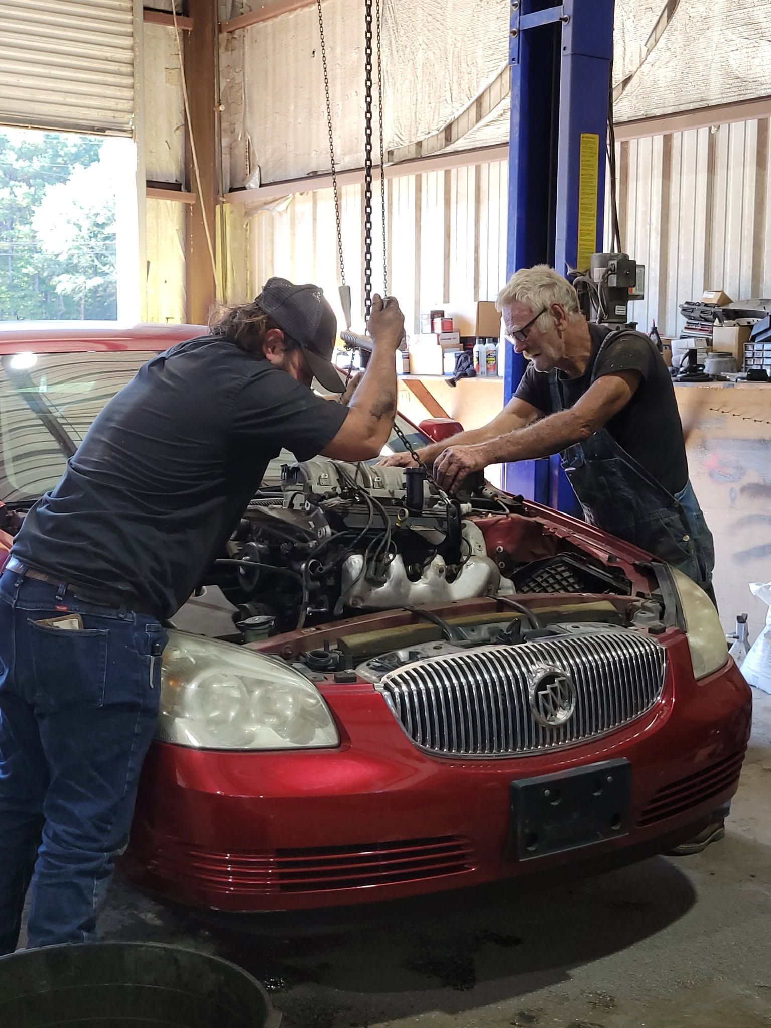Two mechanics leaning over a red car with its hood open inside a workshop