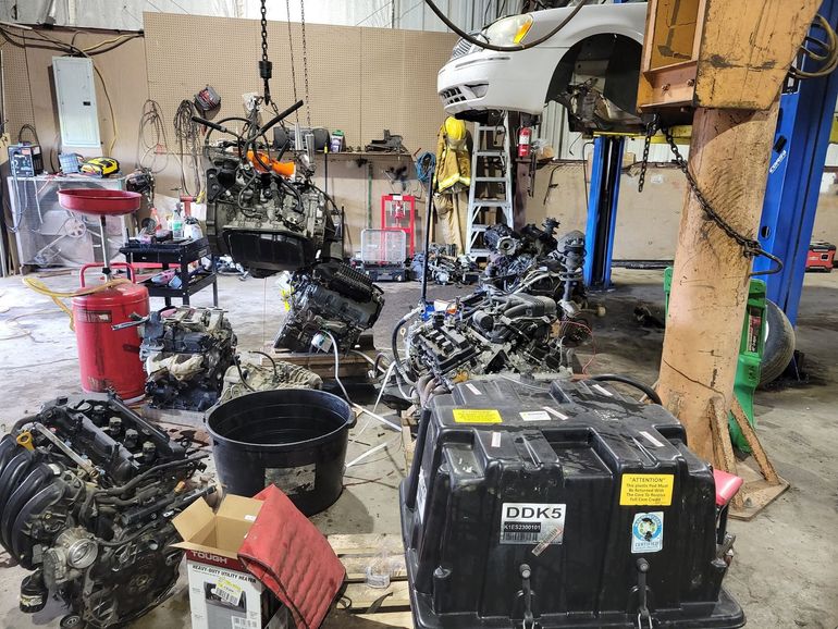 Auto repair shop with several disassembled engines and parts on the floor, under a lifted car.