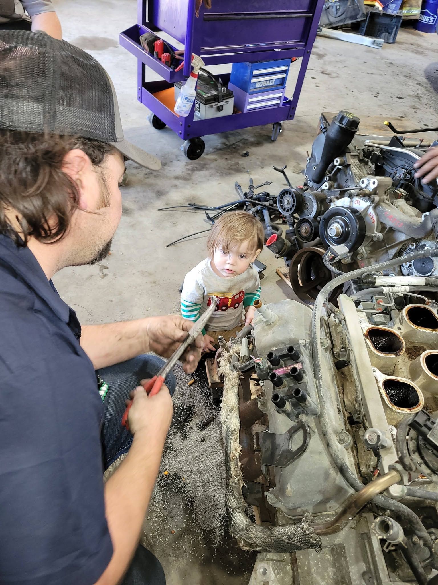 Person crouches beside a toddler near a car engine in a garage, with tools and a purple cart nearby