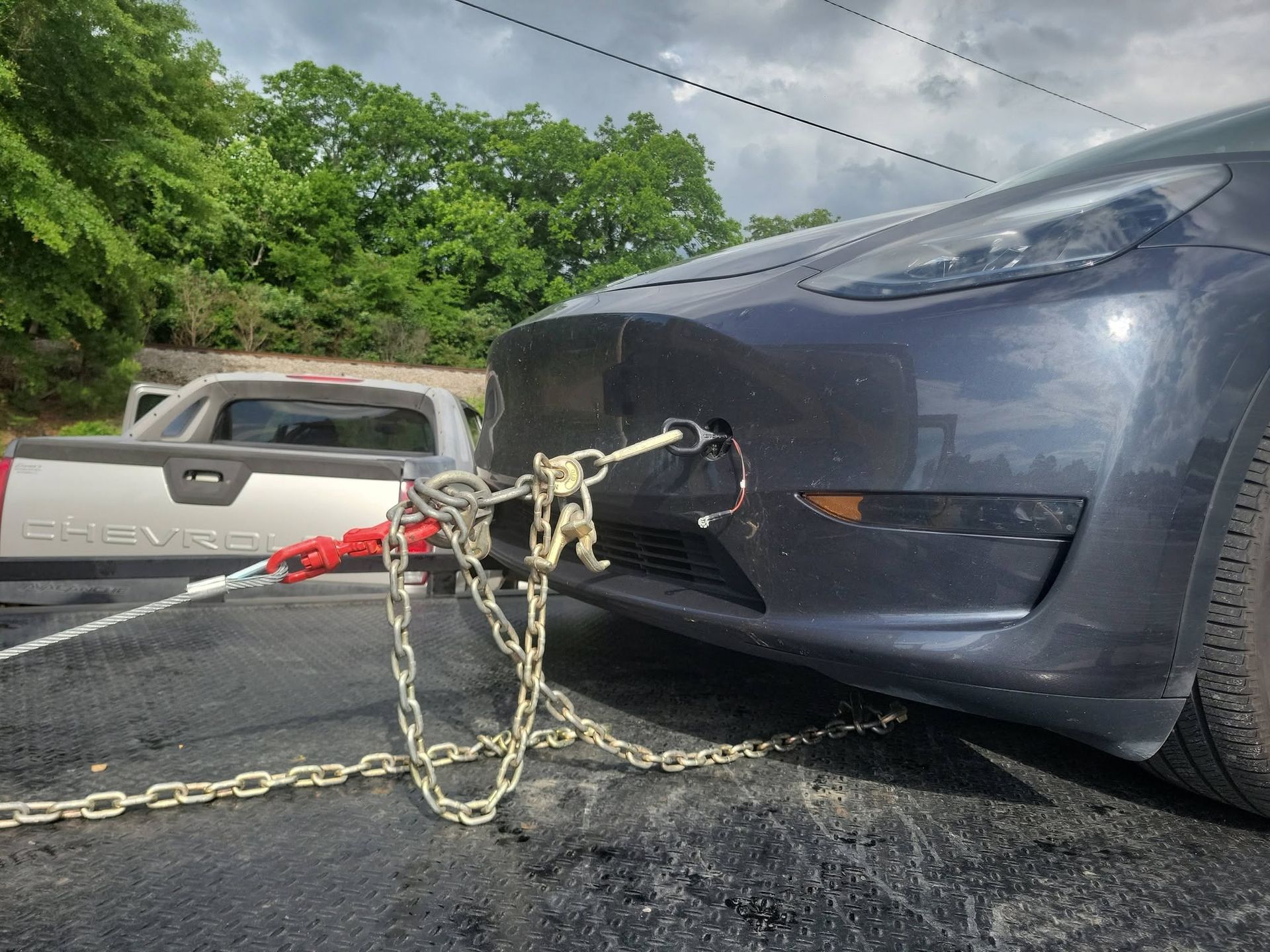 Tow truck chains attached to the front of a dark blue car on a roadside, with another vehicle behind.