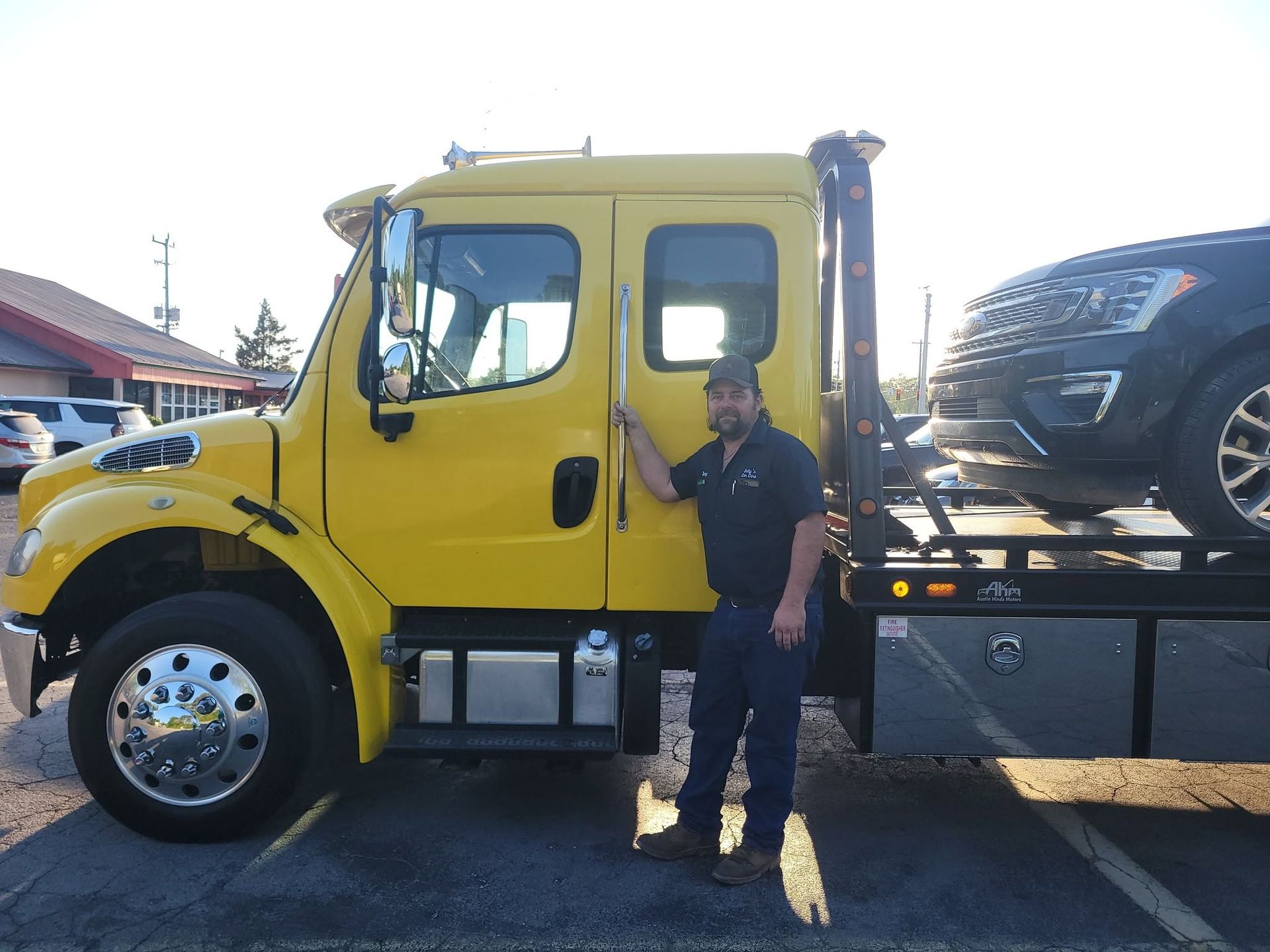 Yellow tow truck with a car on the flatbed and a person standing beside it outdoors.
