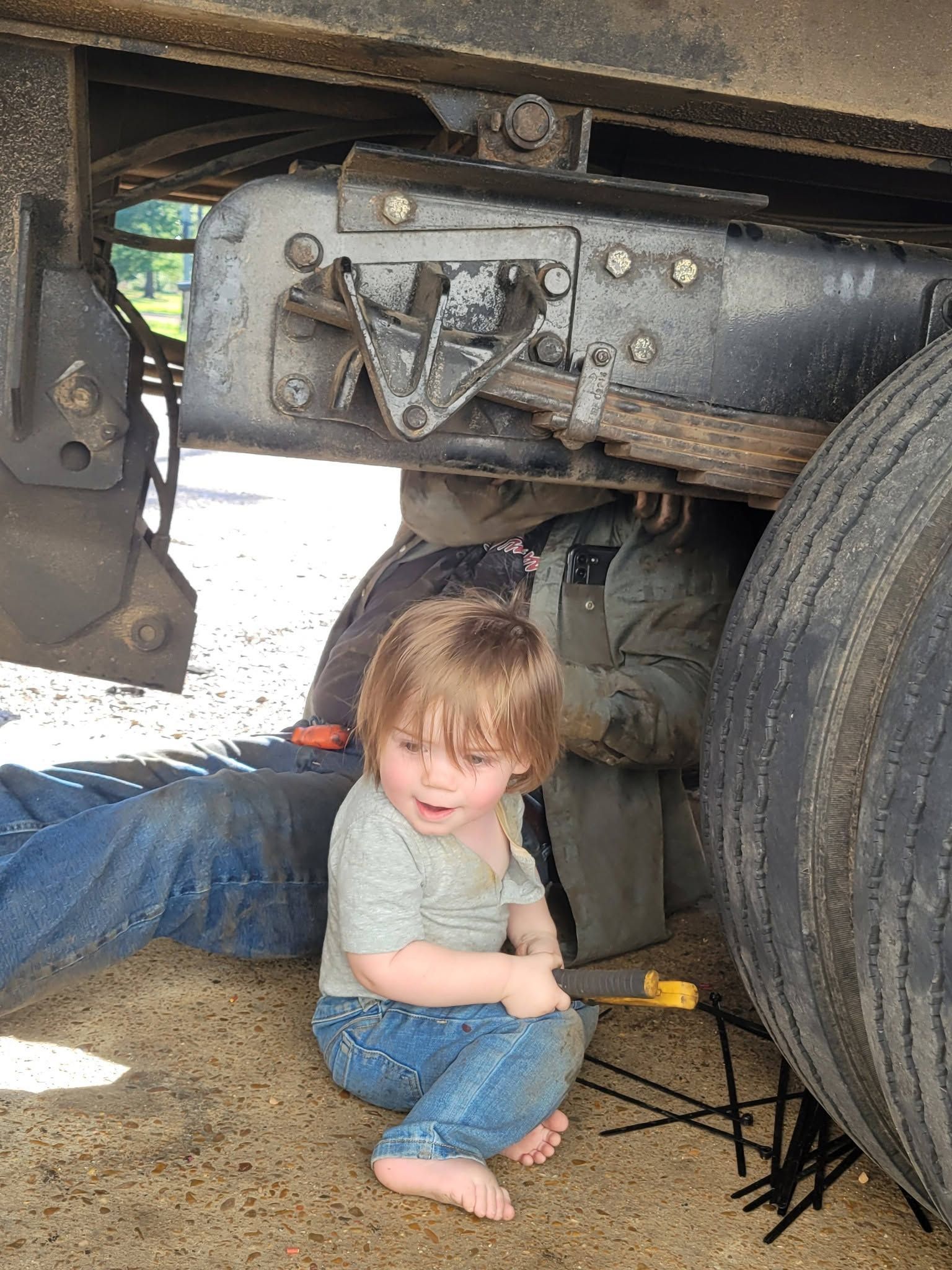 Child sitting under a truck near a large tire, holding a toy wrench, in a garage or repair area