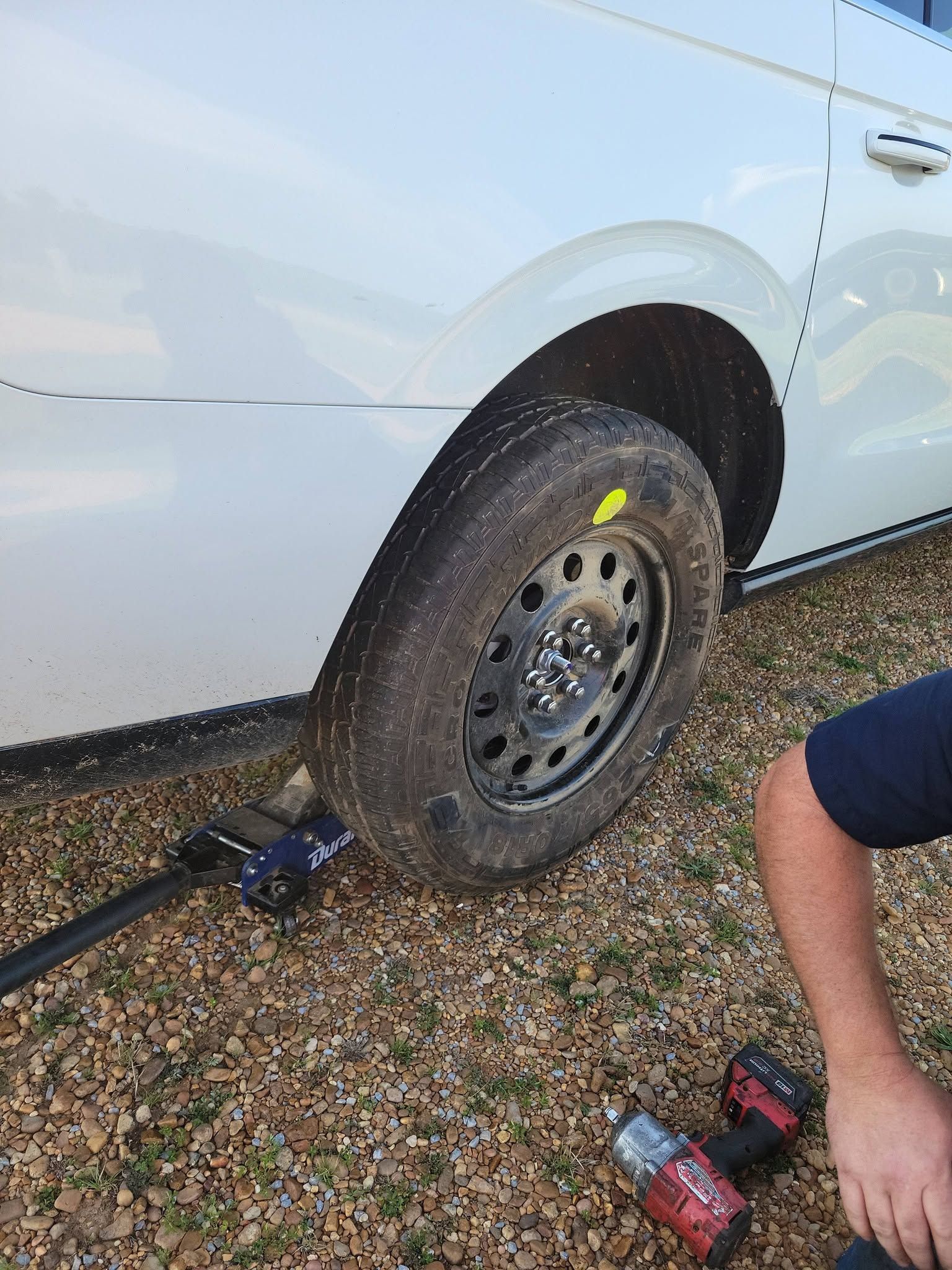 Flat tire on a white car with a jack under the wheel; a person’s arm and tools are visible on gravel.