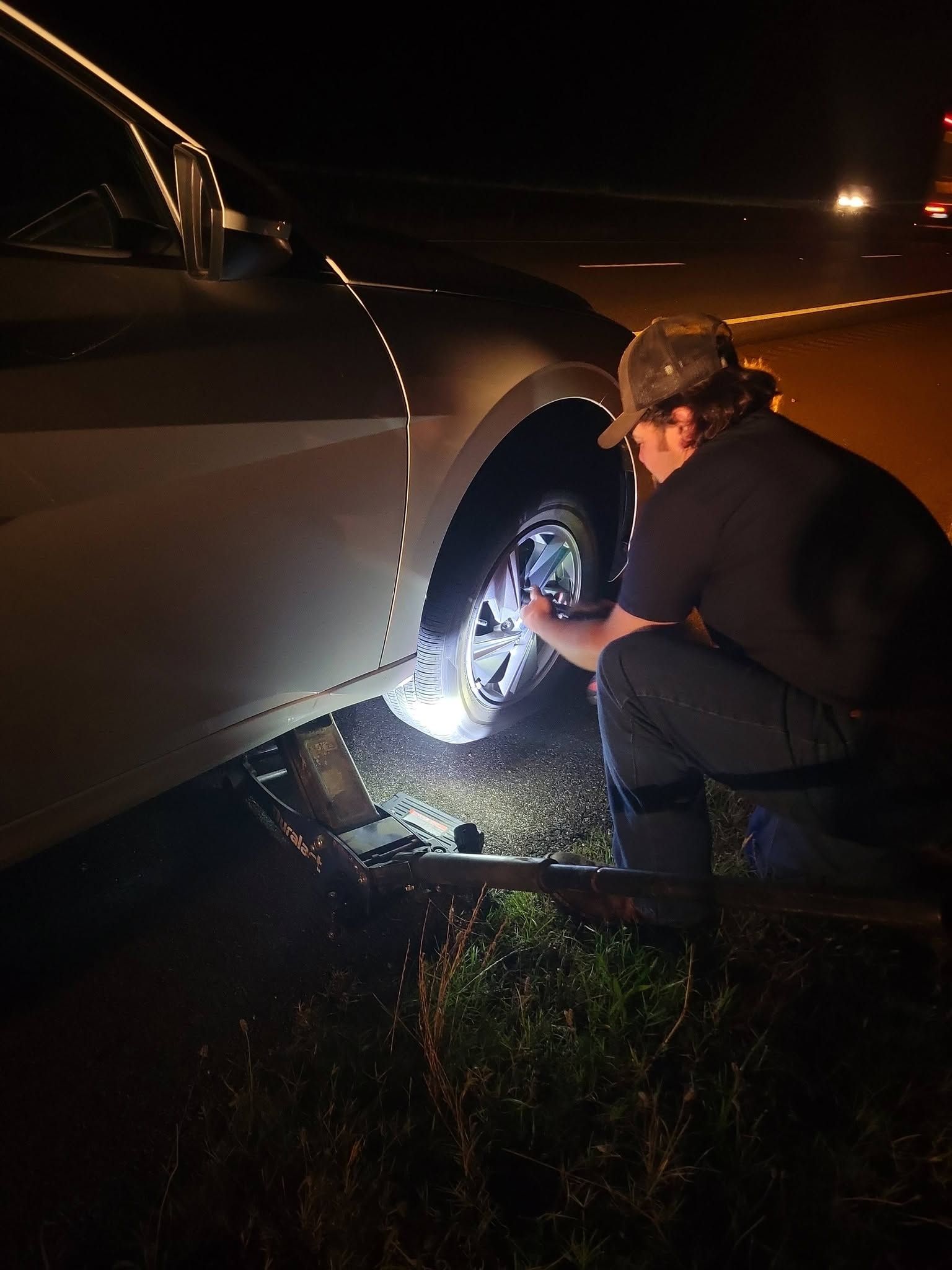 Person kneels by a car wheel at night, using a bright light to inspect or repair a flat tire.