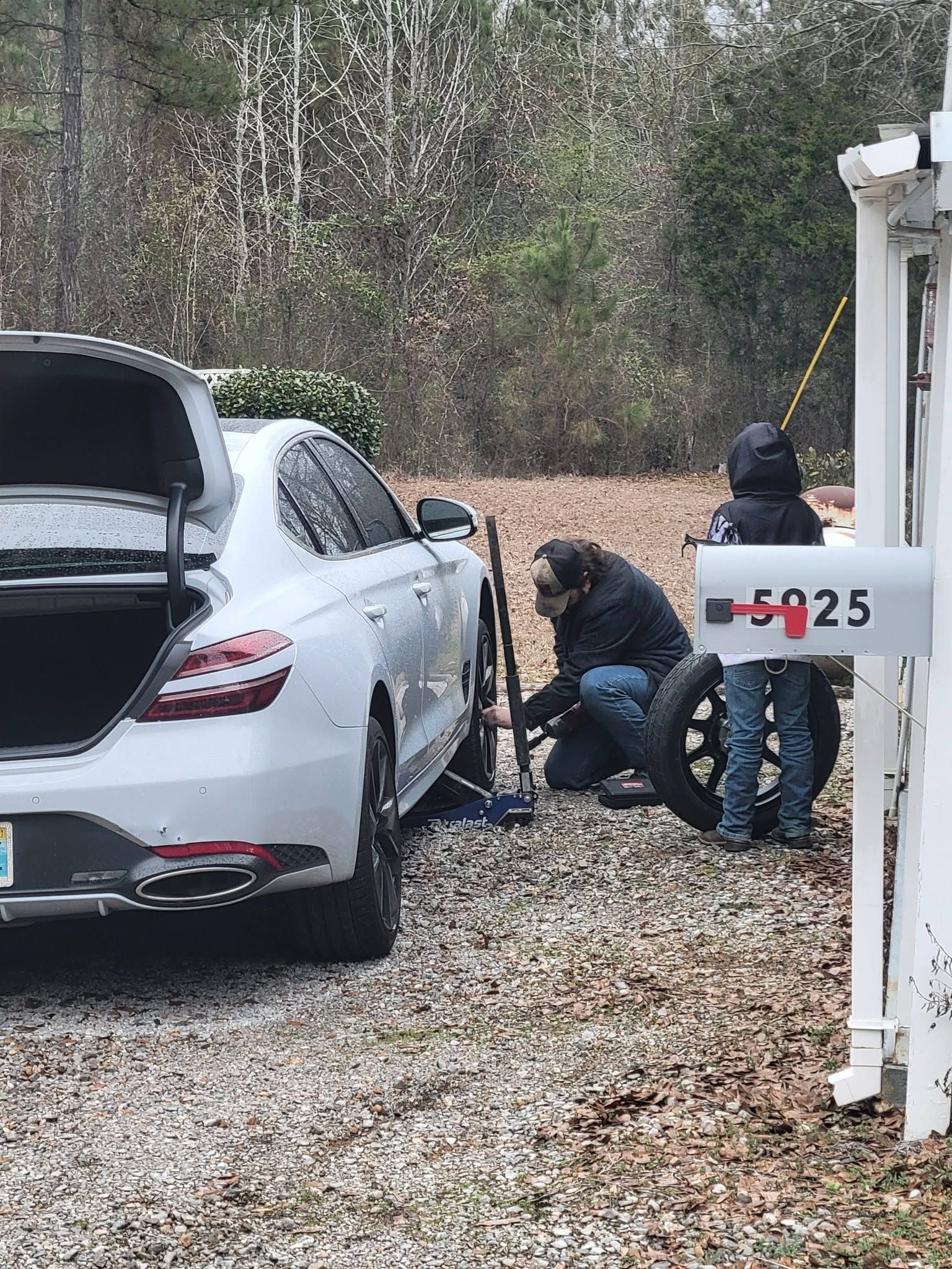 Two people crouch by a white SUV’s front wheel on a gravel driveway with a tow truck nearby.