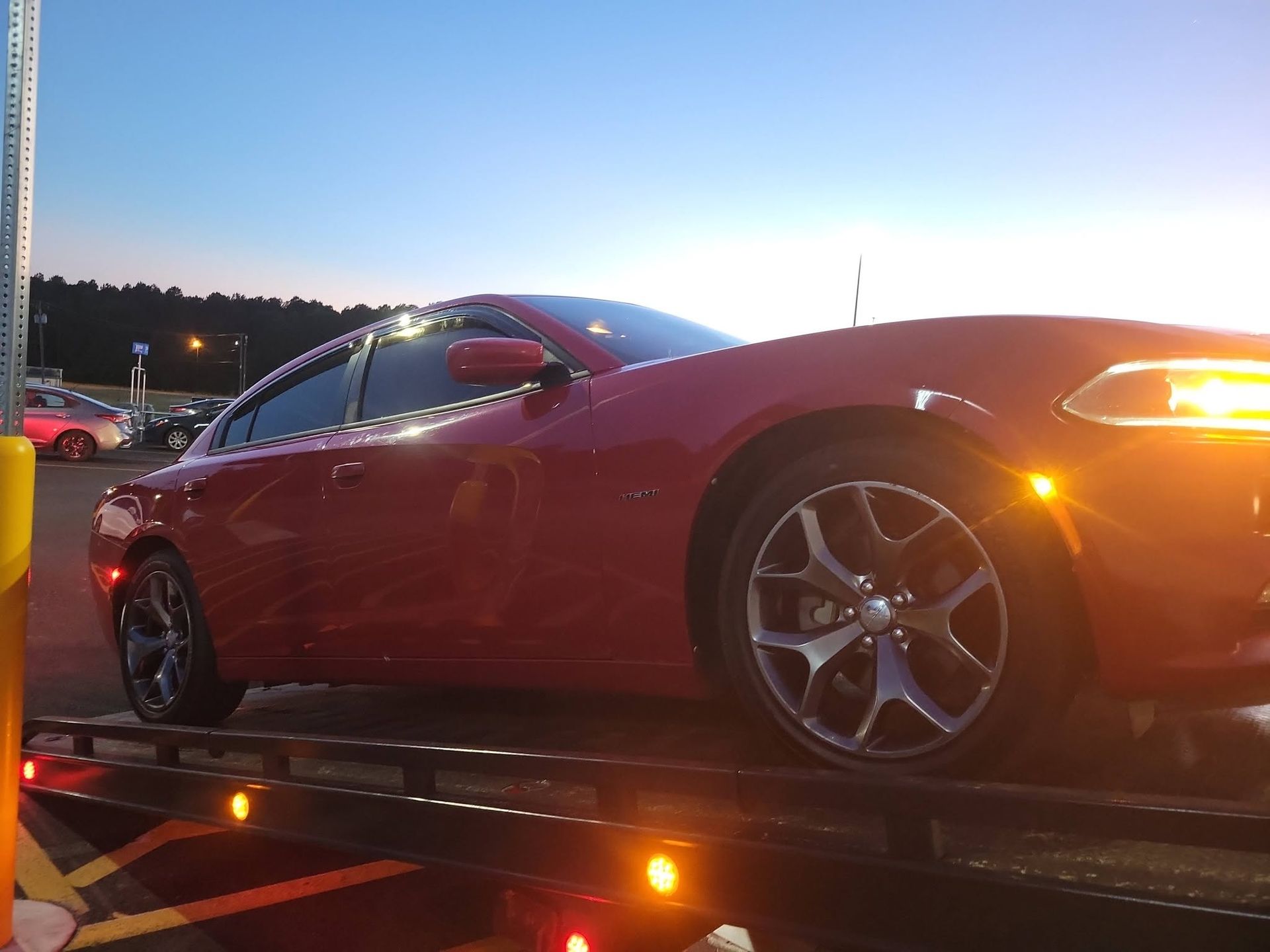 Red sports car on a flatbed tow truck at dusk, headlights on.