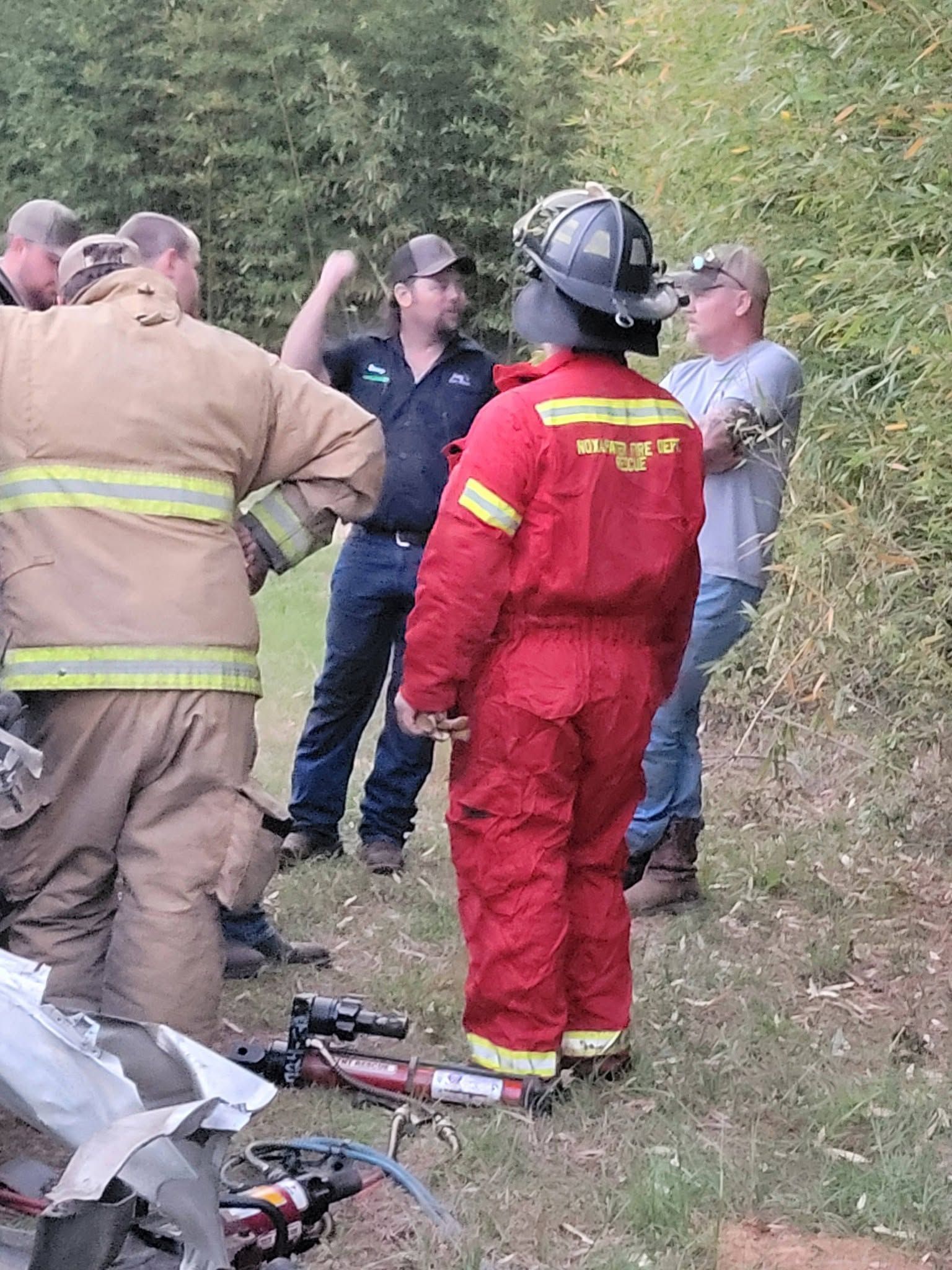 Emergency responders in tan and red uniforms stand by a wooded roadside near rescue equipment.