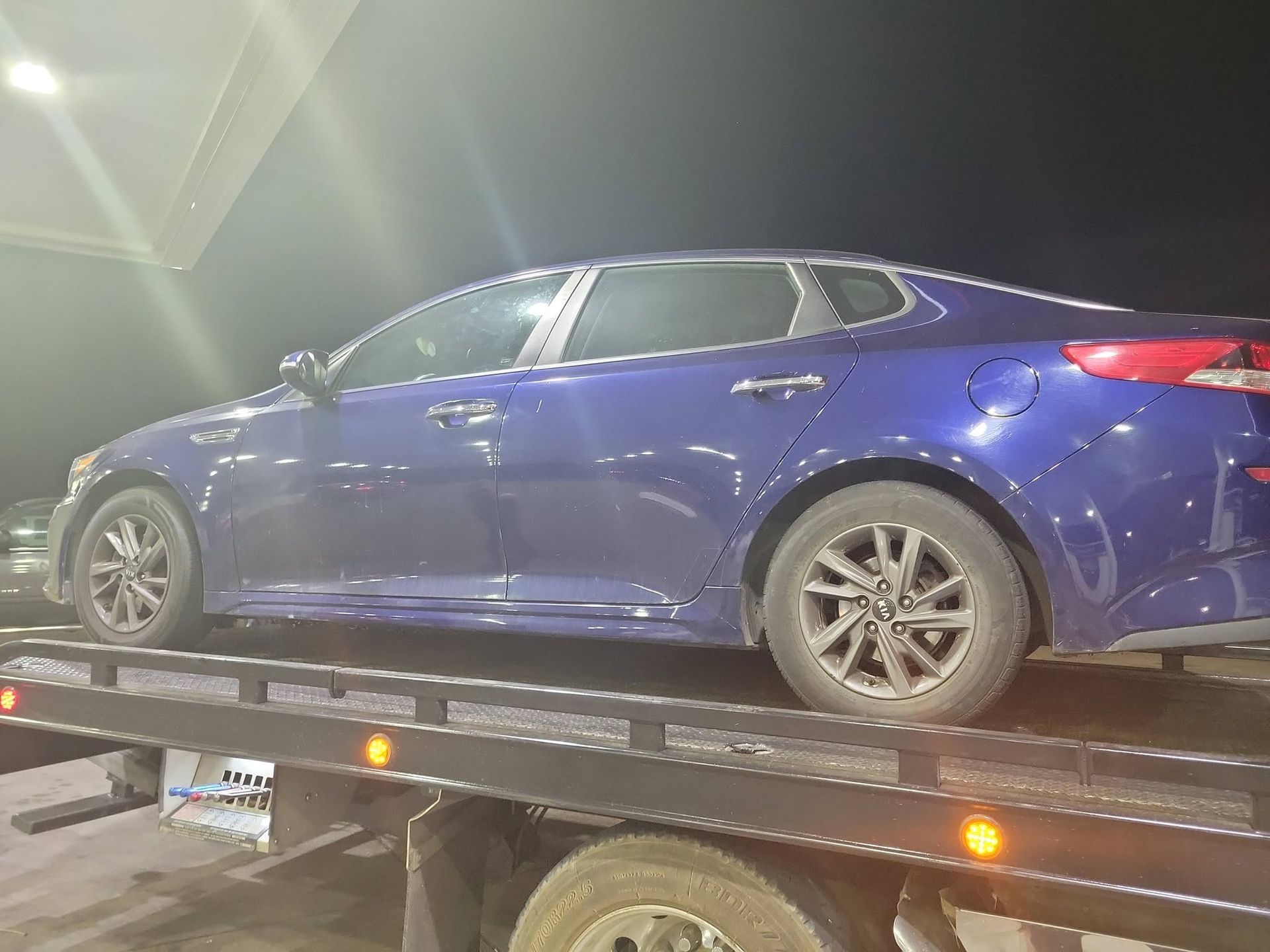 Blue sedan on a tow truck at night, seen from the side with amber lights glowing.
