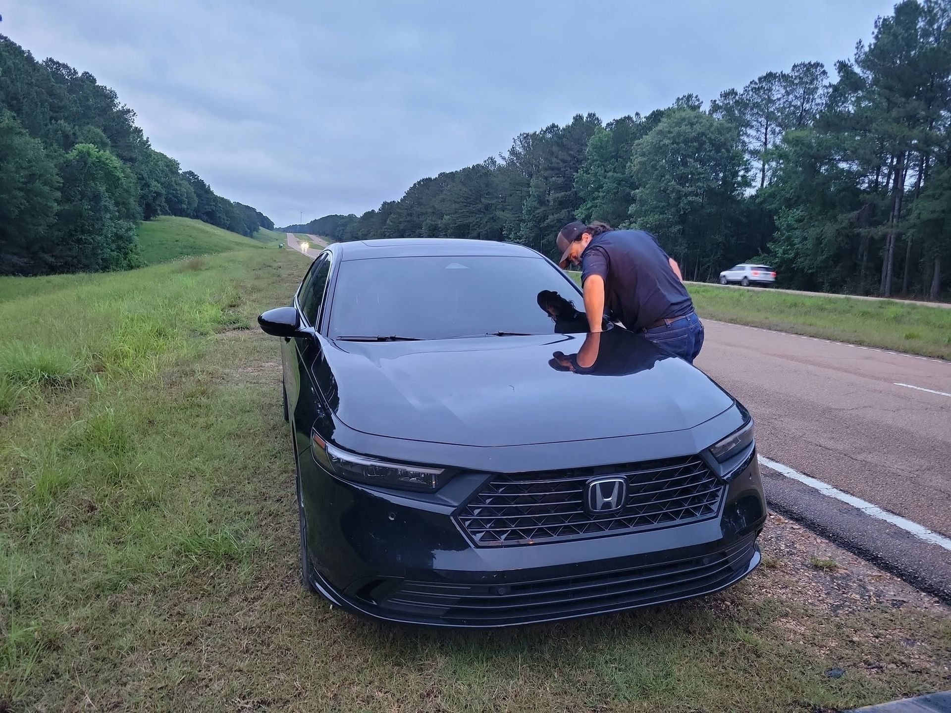 Person leaning over the hood of a black car parked on a roadside shoulder.