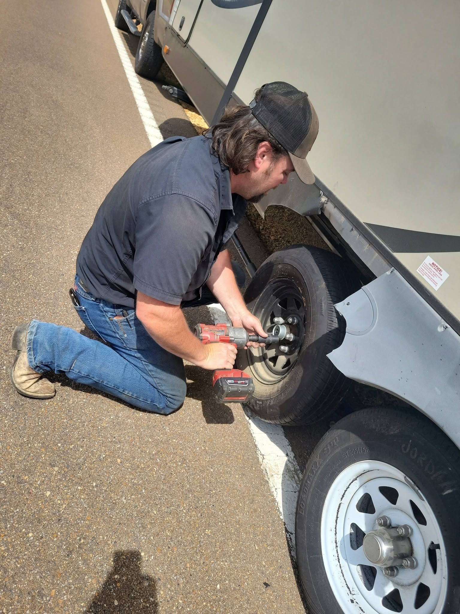 Mechanic kneeling beside a car, using tools to work on a wheel on a roadside.