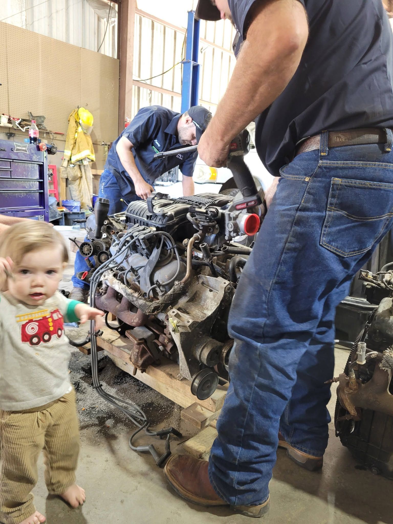 Toddler beside adults working on a muddy engine in a workshop
