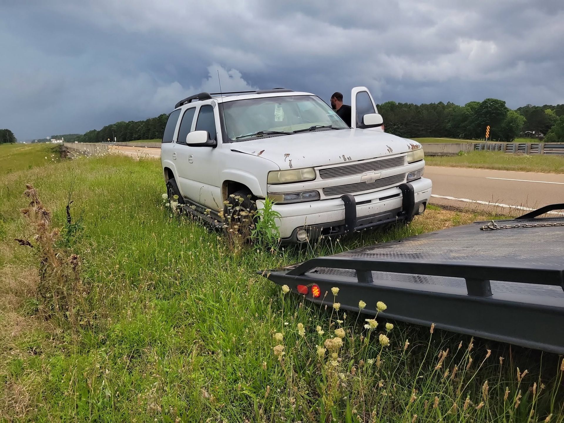 White SUV in roadside ditch beside a tow truck on a grassy highway shoulder under cloudy skies