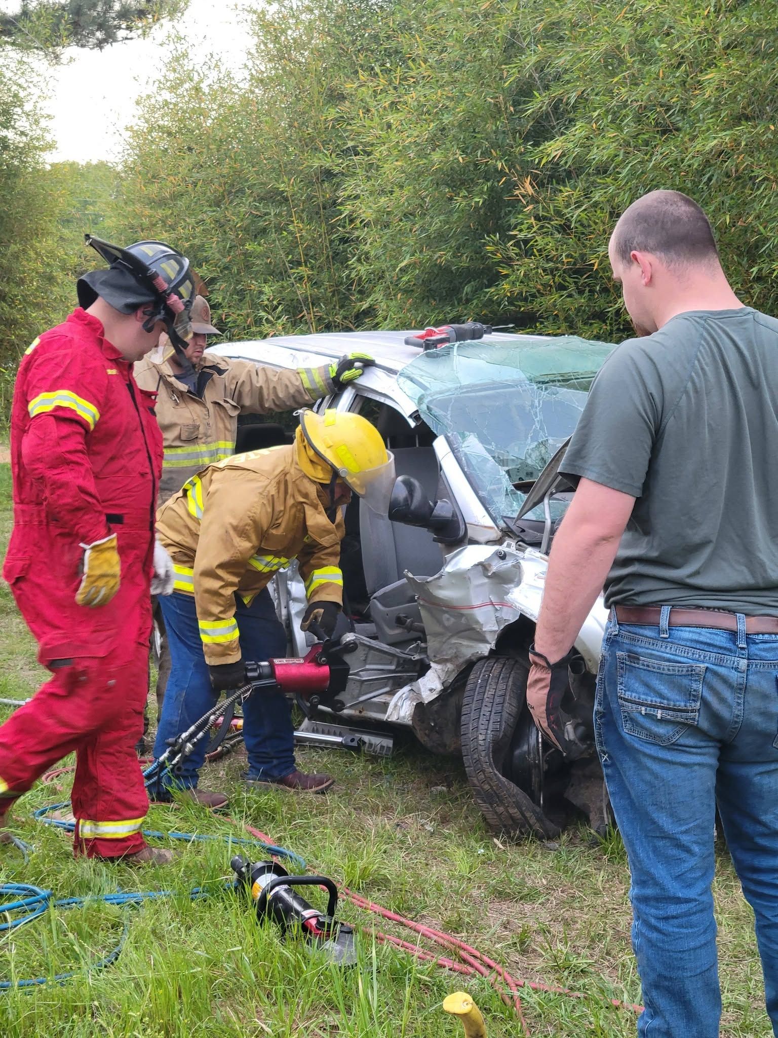 Rescue workers in red and yellow gear assess a crashed silver car on a grassy roadside.