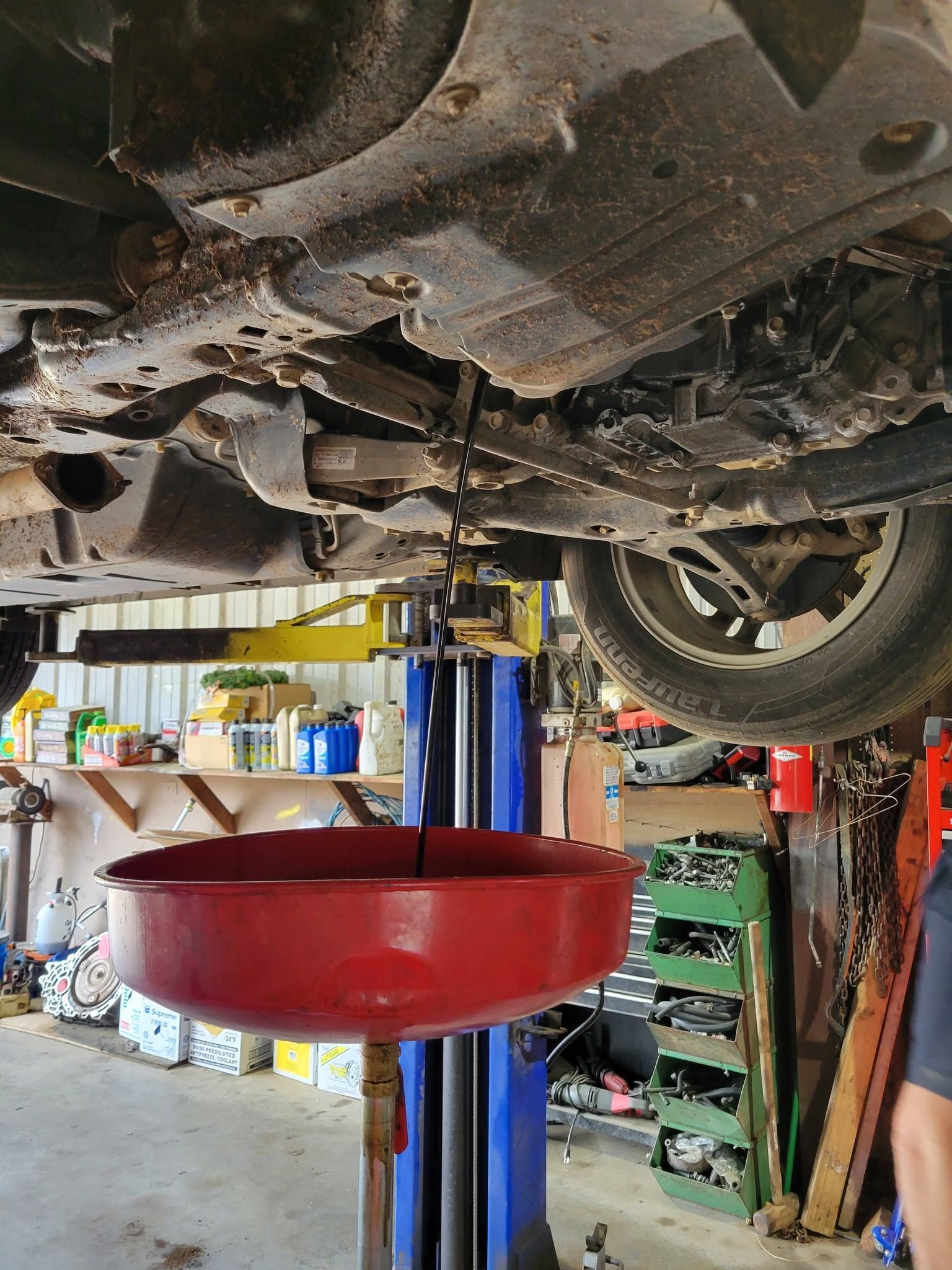 Car underside on a lift in a repair shop, with a red oil drain pan beneath it.